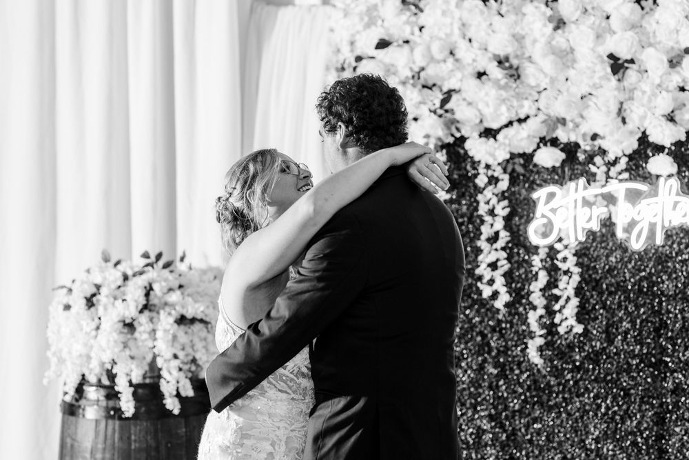 Two people embrace during a wedding celebration with elegant white floral arrangements in the background.