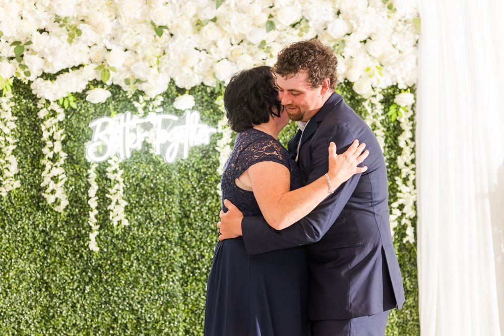 Couple embraces during tender moment under white floral arch and greenery wall backdrop.
