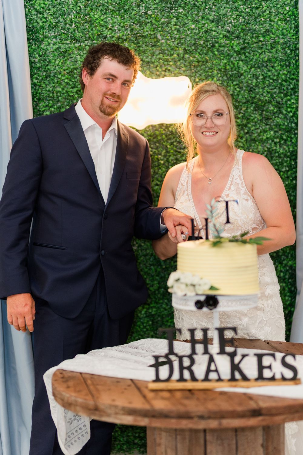 Couple in formal wedding attire cuts their rustic white wedding cake together on a wooden stand against green hedge backdrop.
