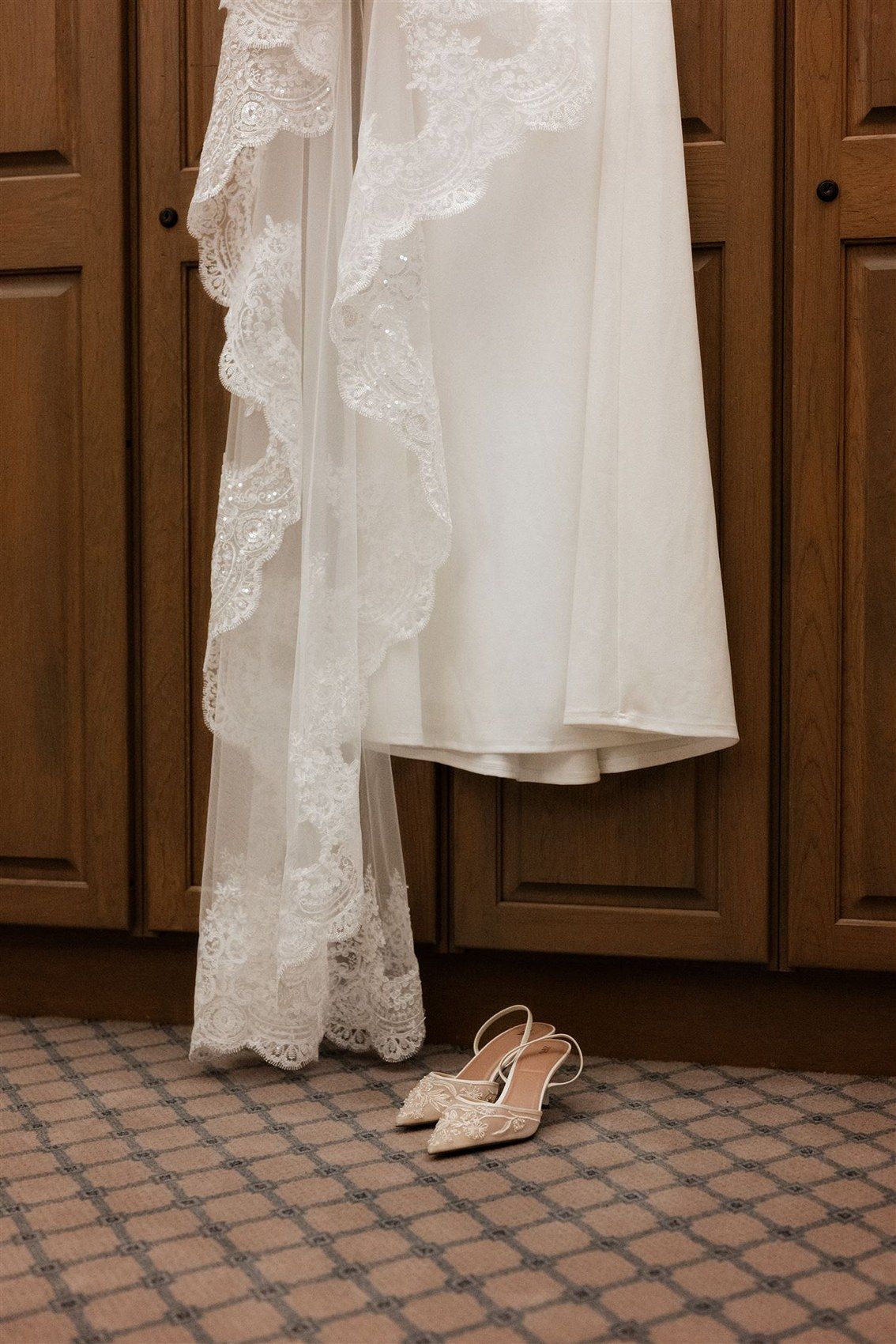 A white lace wedding dress hanging against wooden doors with delicate bridal shoes placed on a tiled floor below.