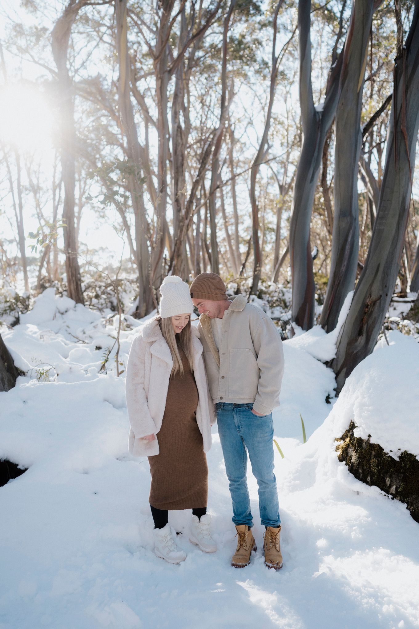 A couple in warm winter clothing standing together on a snow-covered forest path with sunlit eucalyptus trees in background.