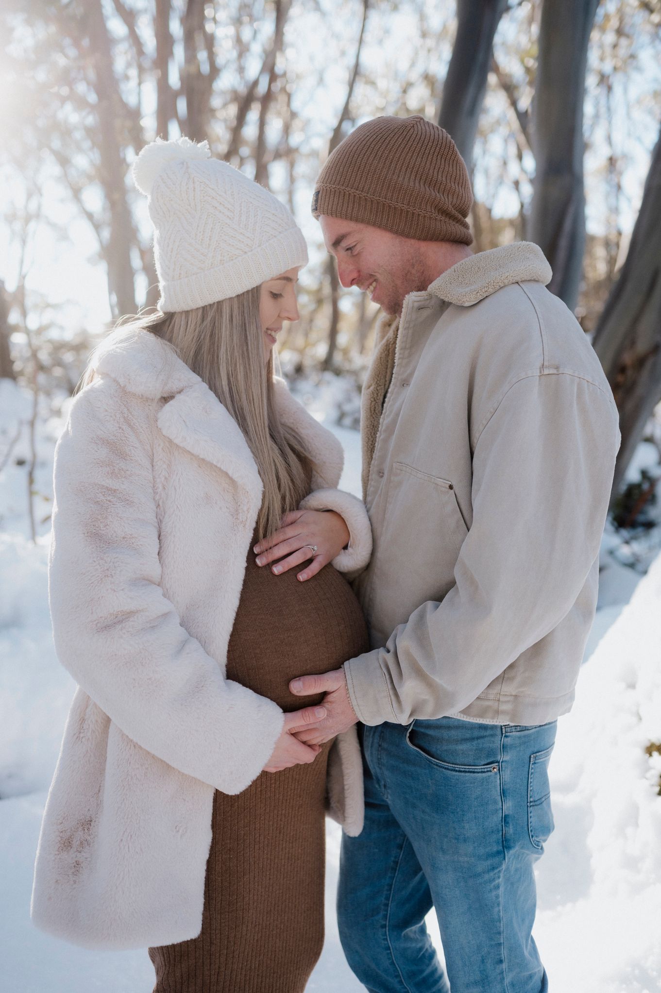 A couple in winter coats share an intimate moment in the snow, with the woman wearing a brown dress and white beanie.