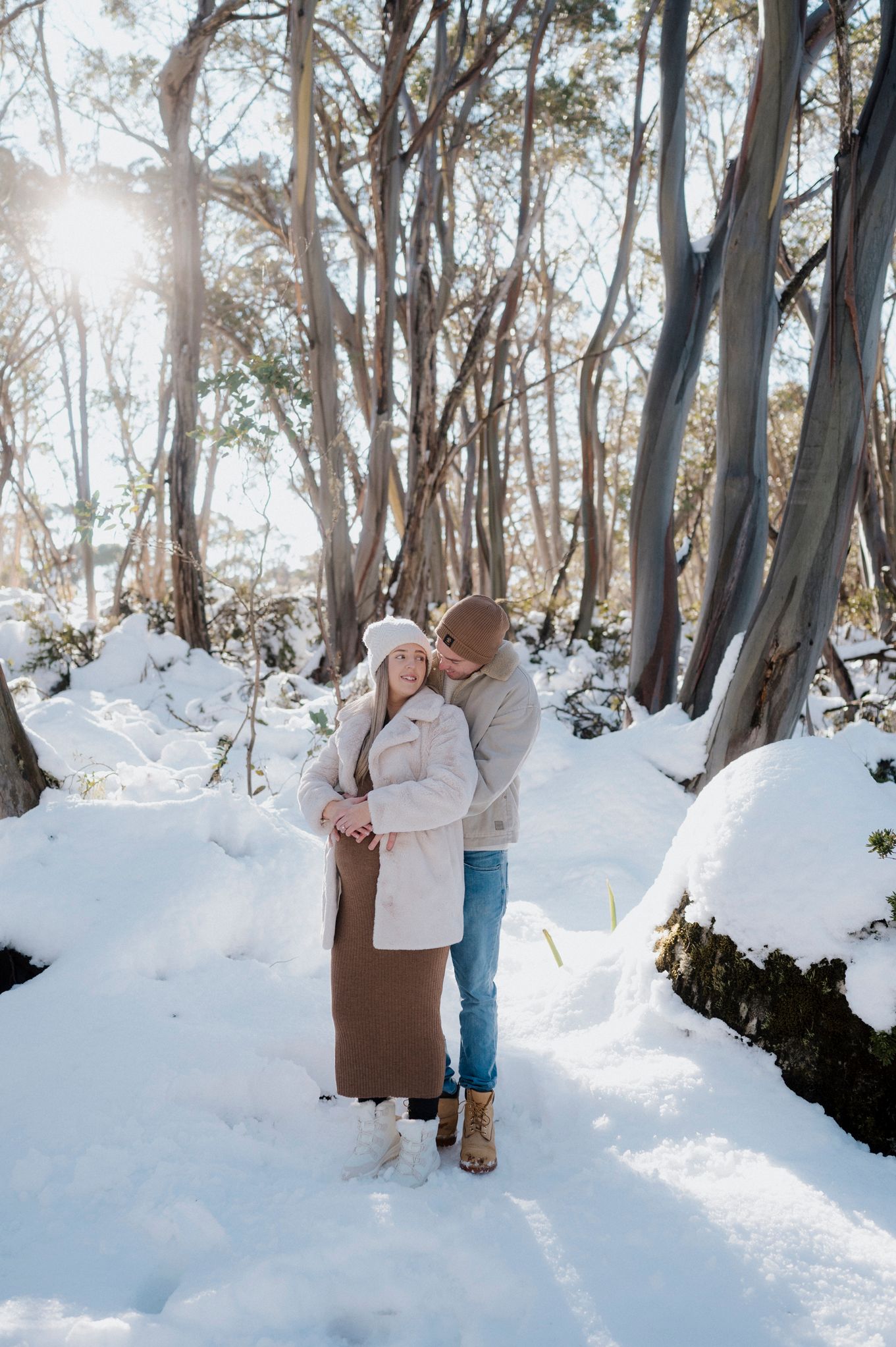 A couple embraces while standing in a snowy forest with sunlight streaming through tall eucalyptus trees.