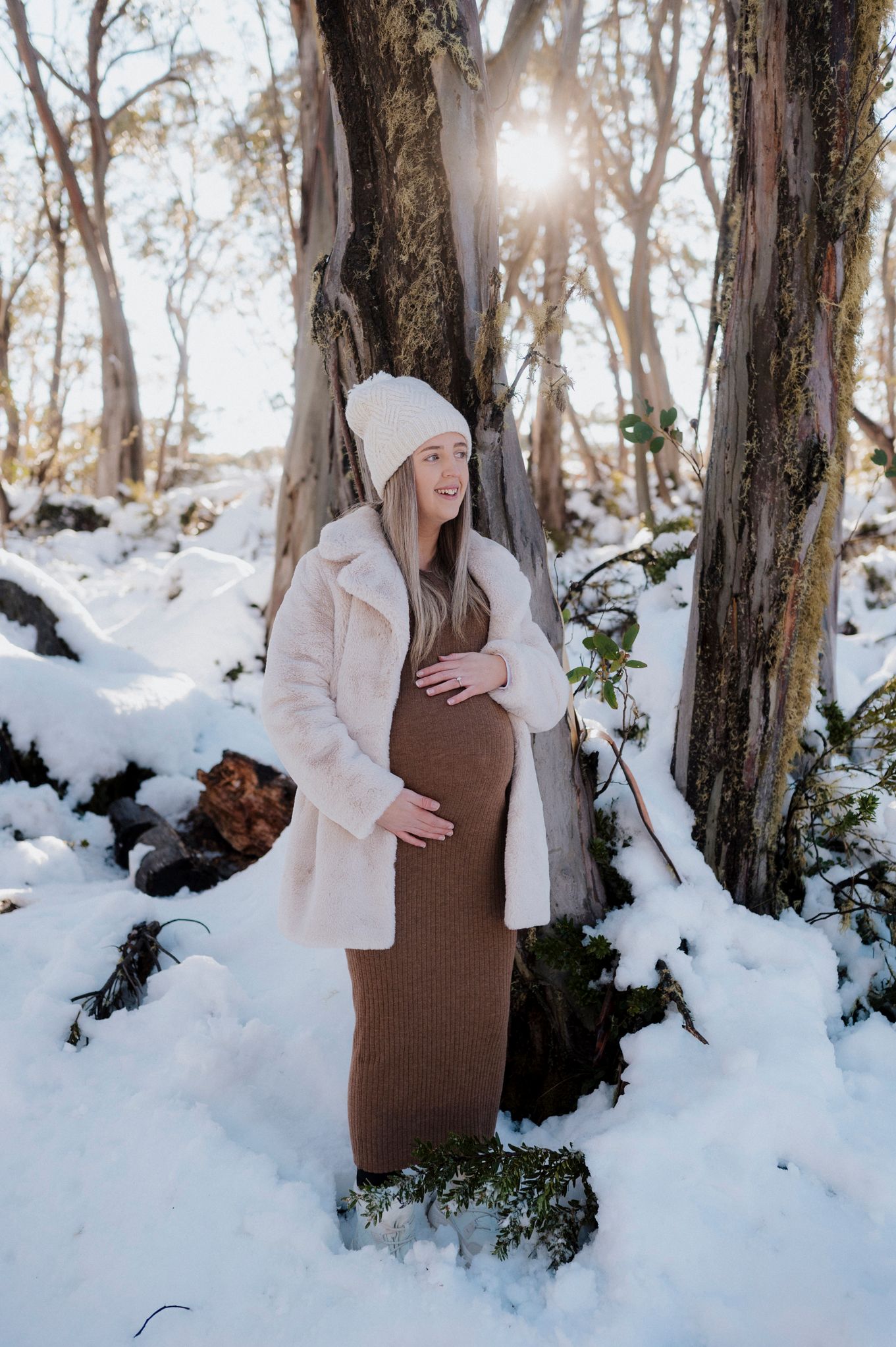 Winter maternity photo in a snowy forest with sunlight peeking through trees as someone poses in a brown dress and white coat.