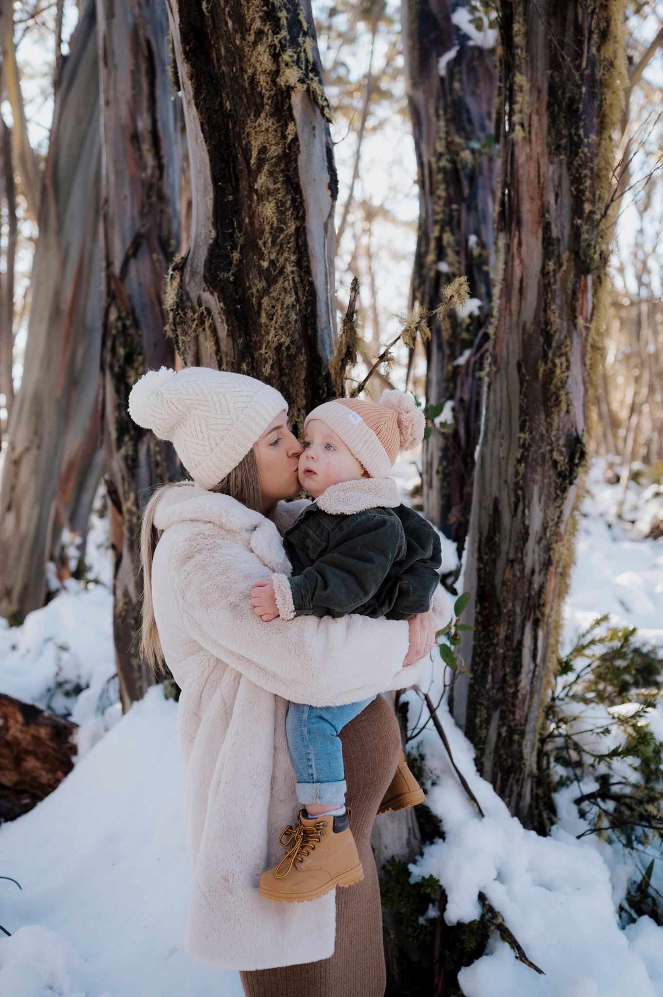 Winter outdoor scene in snowy woods with two people embracing near dark tree trunks in soft natural lighting.