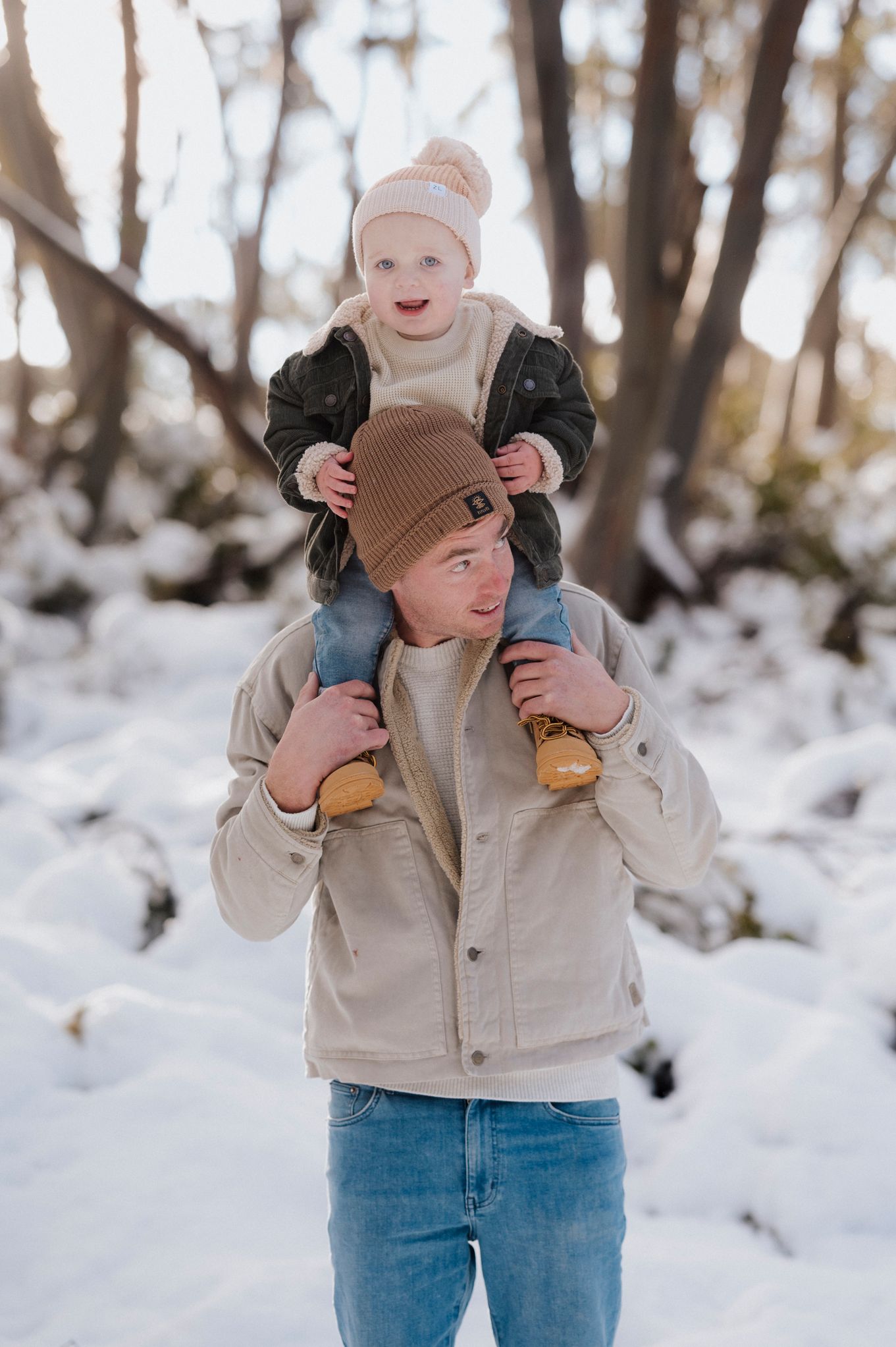 A person in a beige jacket laughs while holding up their child in a snowy winter forest setting.