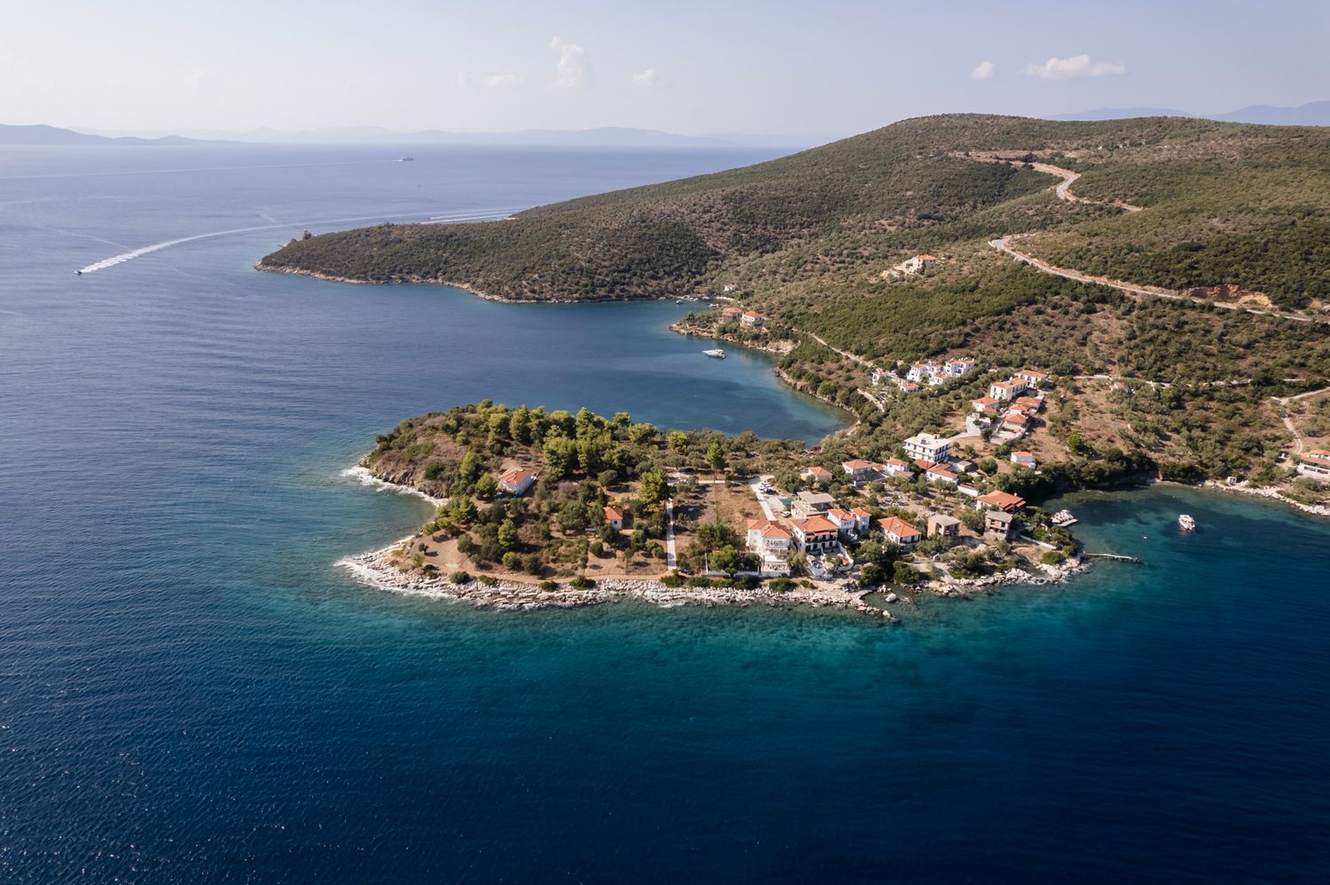 Aerial panoramic view of a coastal Mediterranean town with red roofs nestled along turquoise bay waters.