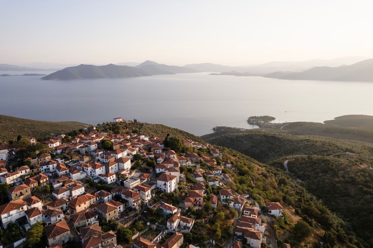 Aerial panoramic view of coastal Mediterranean town with red rooftops nestled on hillside overlooking blue sea and islands.