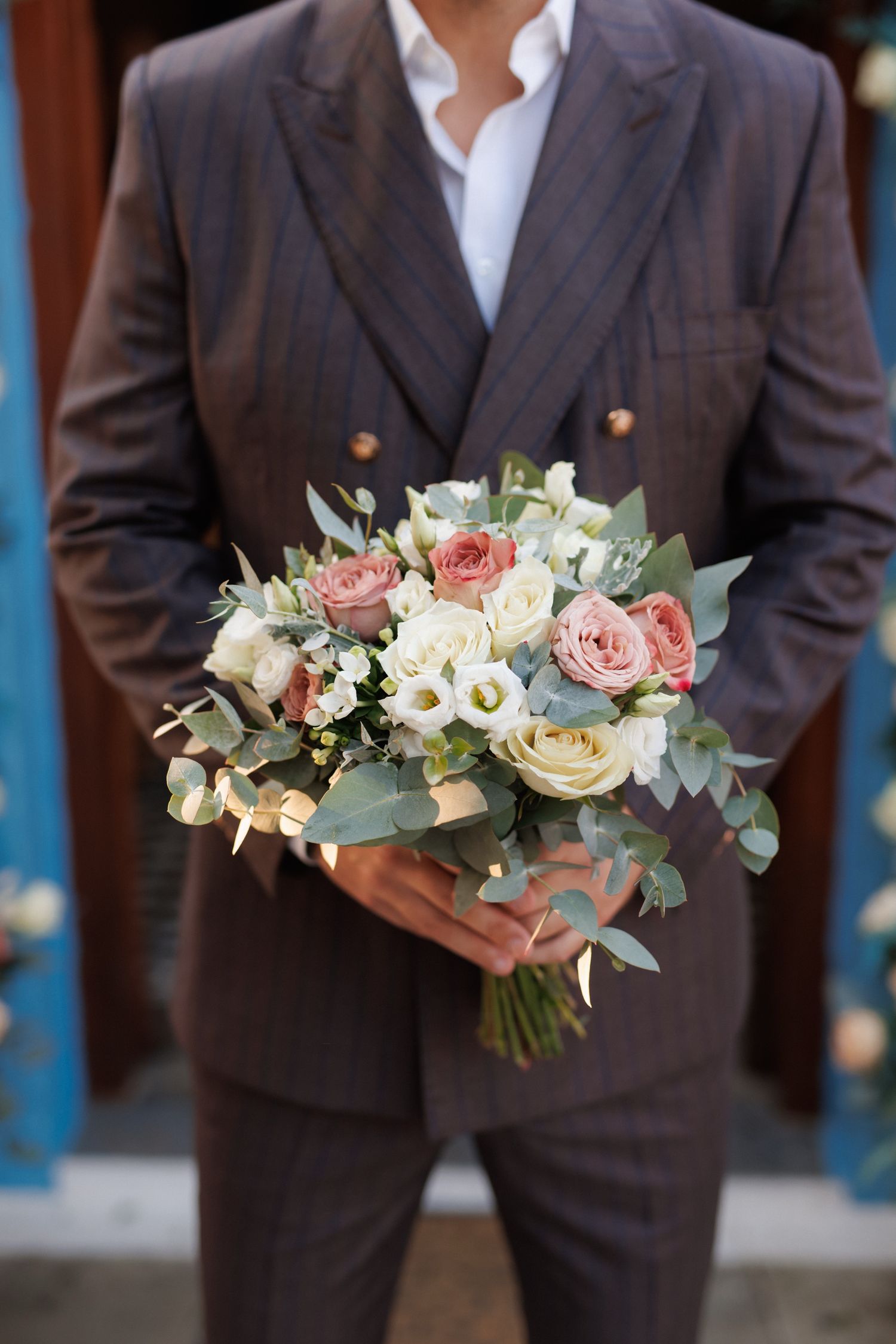 Groomsmen in brown suits hold elegant bouquets of pink and white roses with eucalyptus against a blue backdrop.