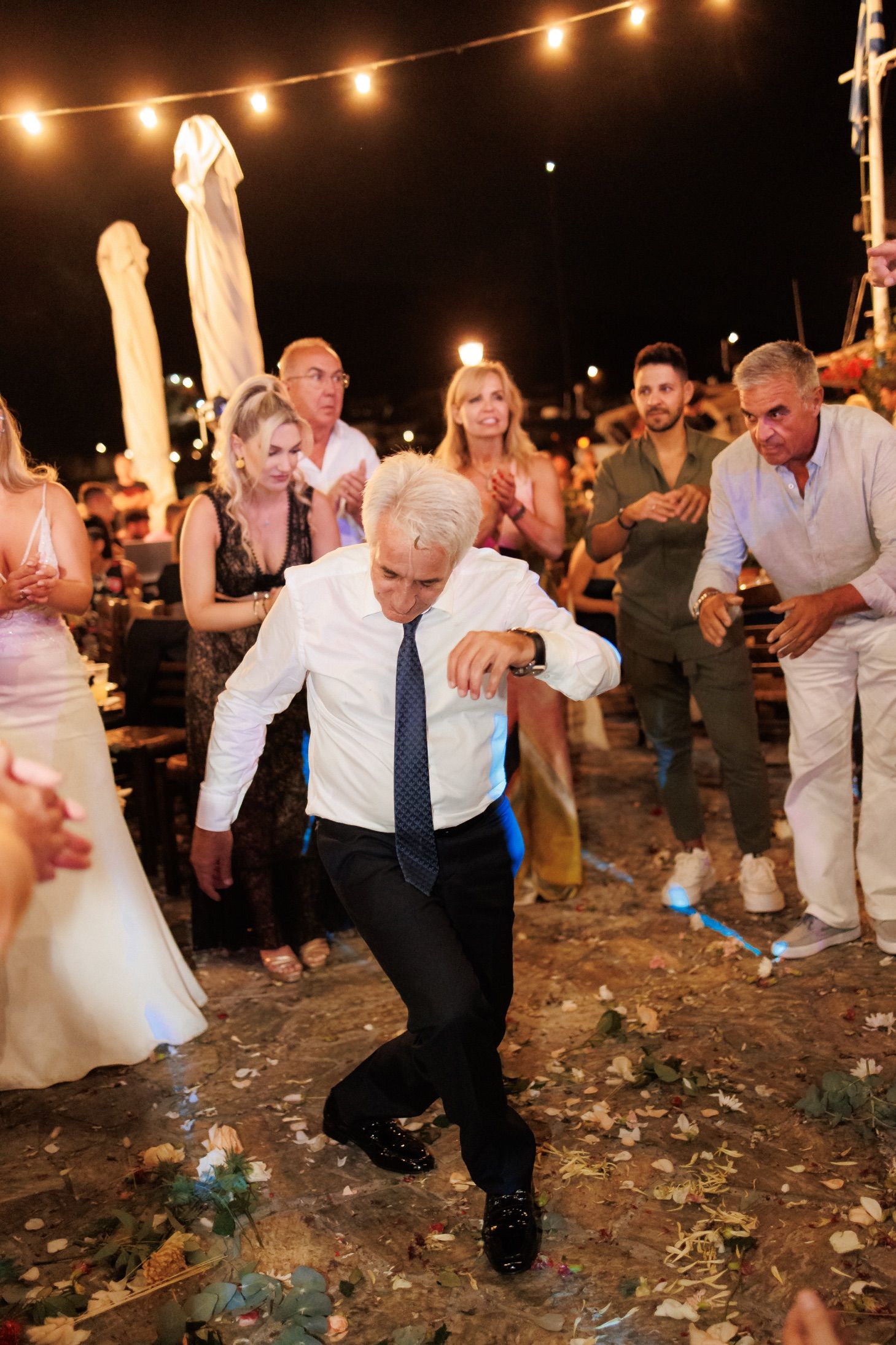 People dancing and celebrating at a nighttime outdoor party with string lights and confetti on the ground.