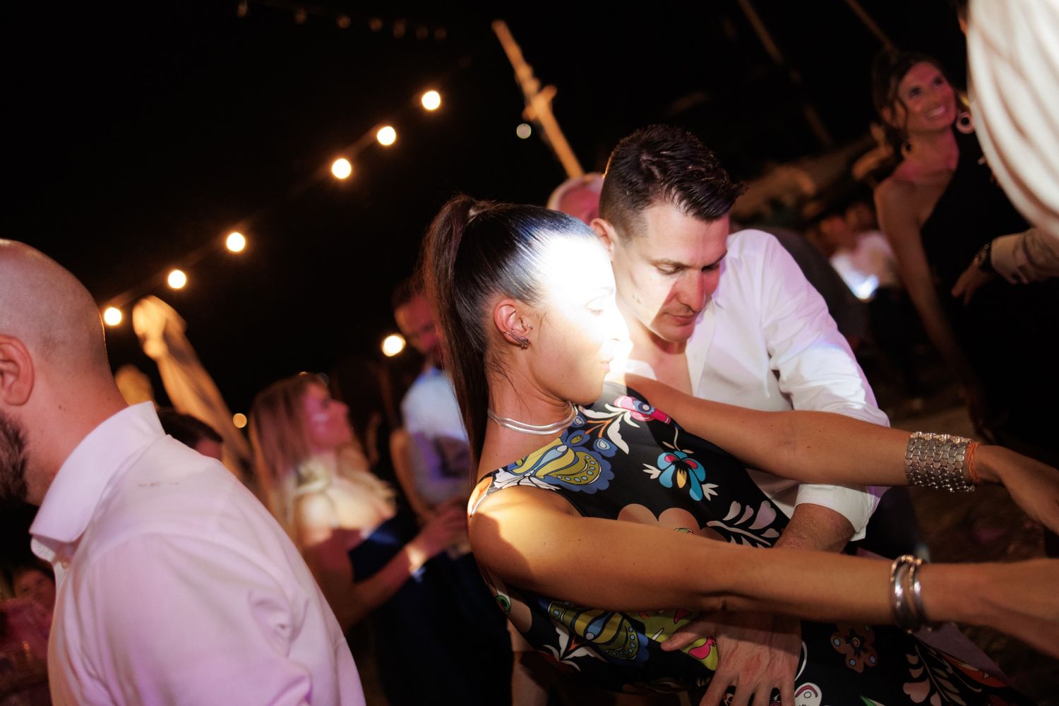 People dancing and celebrating at a nighttime party with string lights illuminating the festive atmosphere.