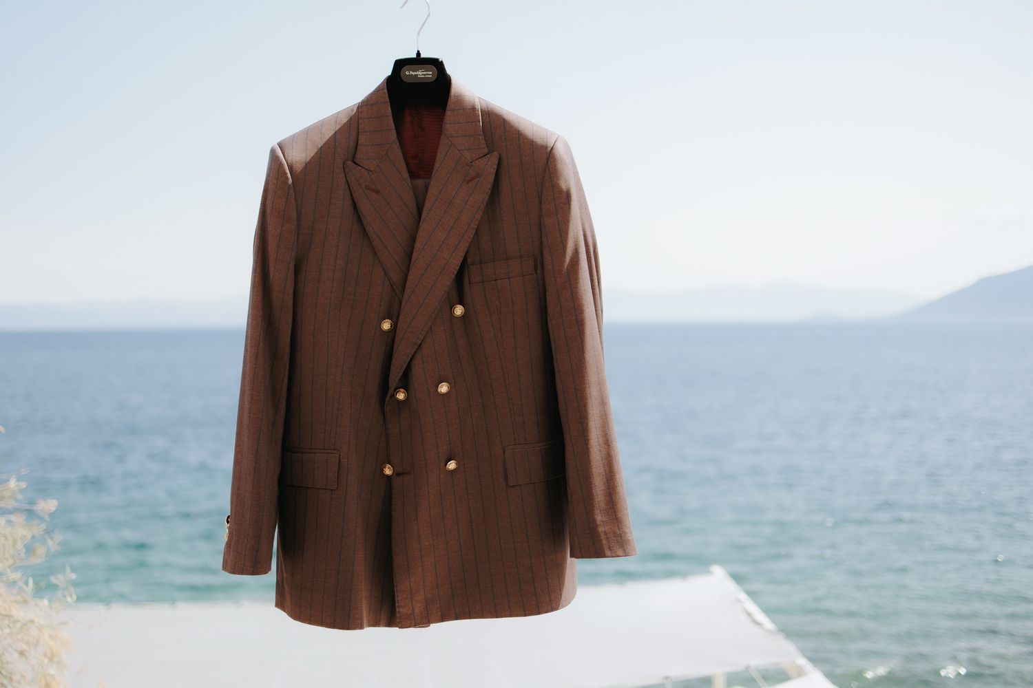 A brown double-breasted suit jacket hangs against a scenic ocean and mountain backdrop.