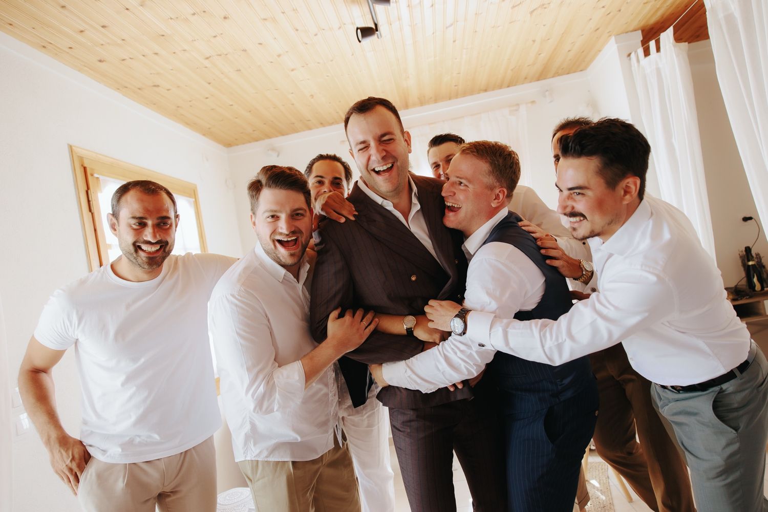 Joyful wedding party members in white shirts and gray pants laughing together in a bright indoor setting.