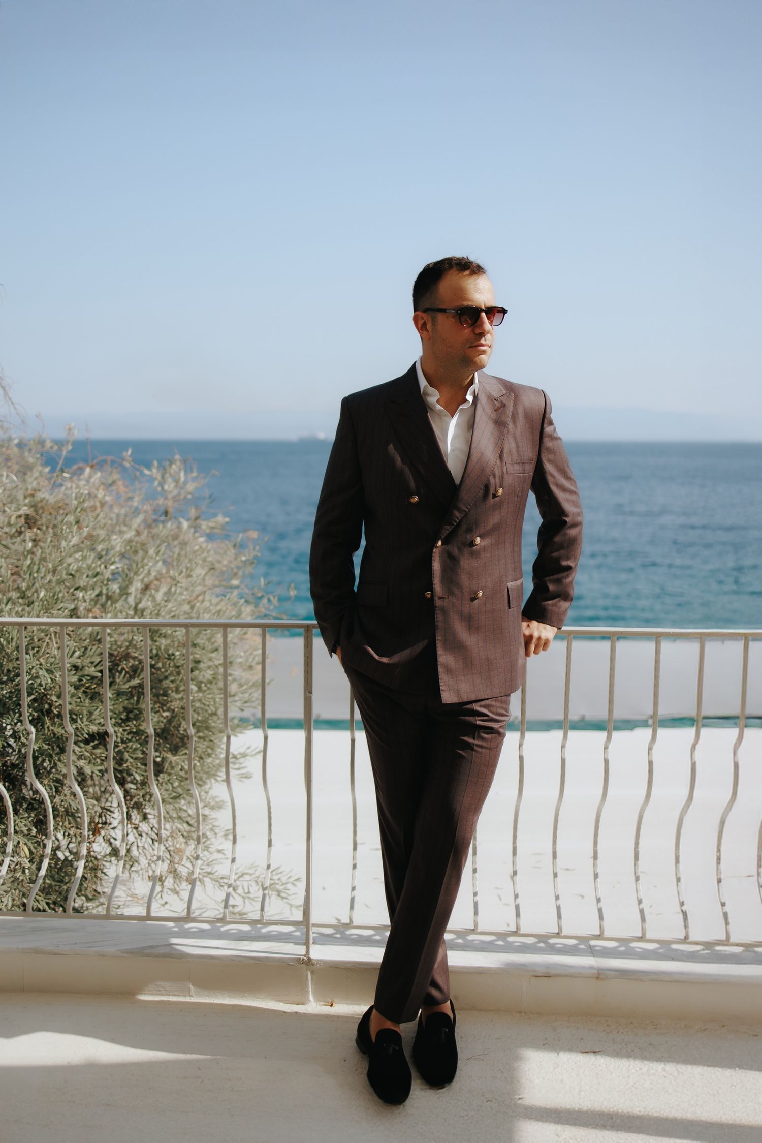 Fashionable person in brown suit poses against ocean balcony view in Mediterranean setting.
