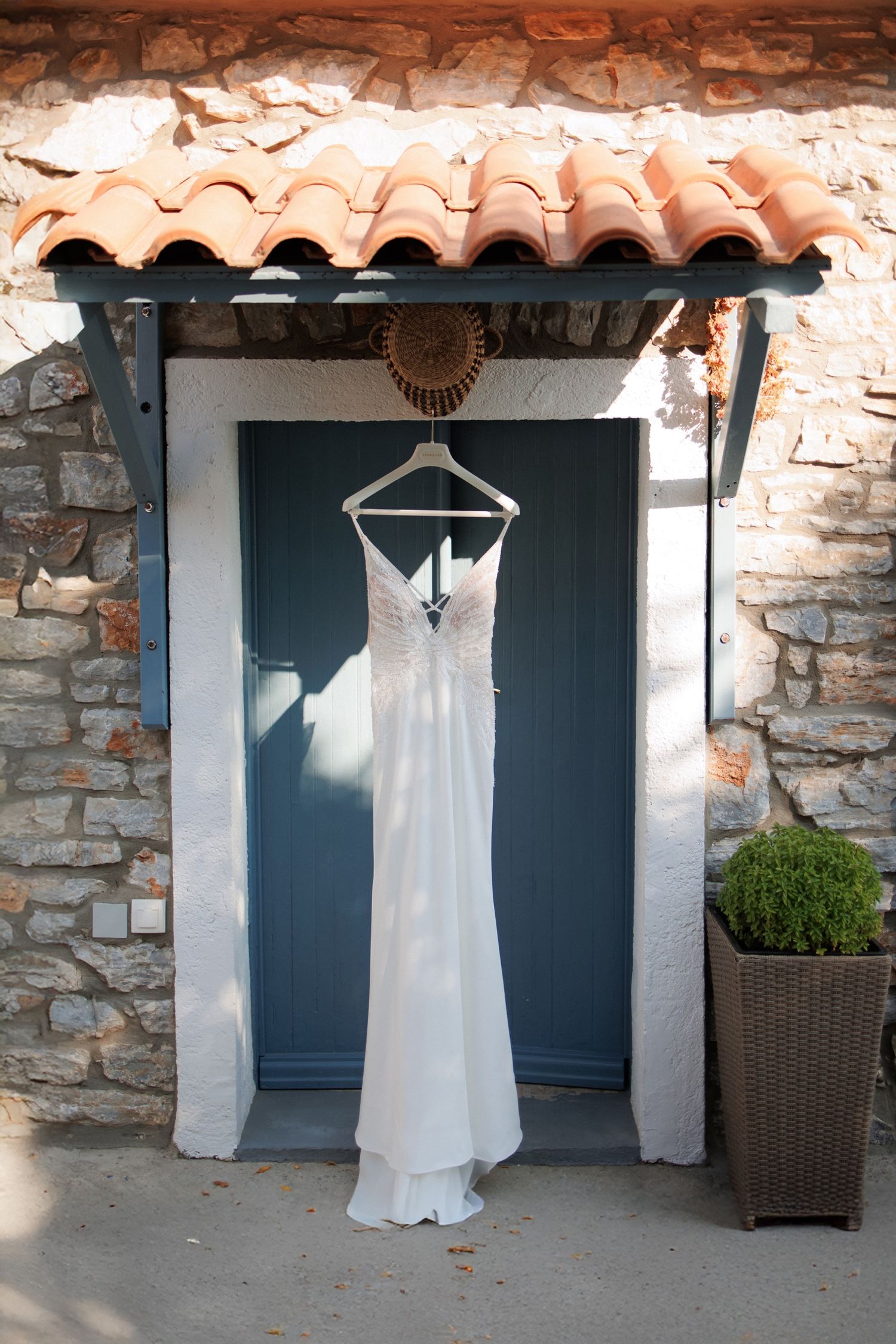 A white wedding dress hangs on a hanger against a blue door with rustic stone walls and terra cotta roof tiles above.