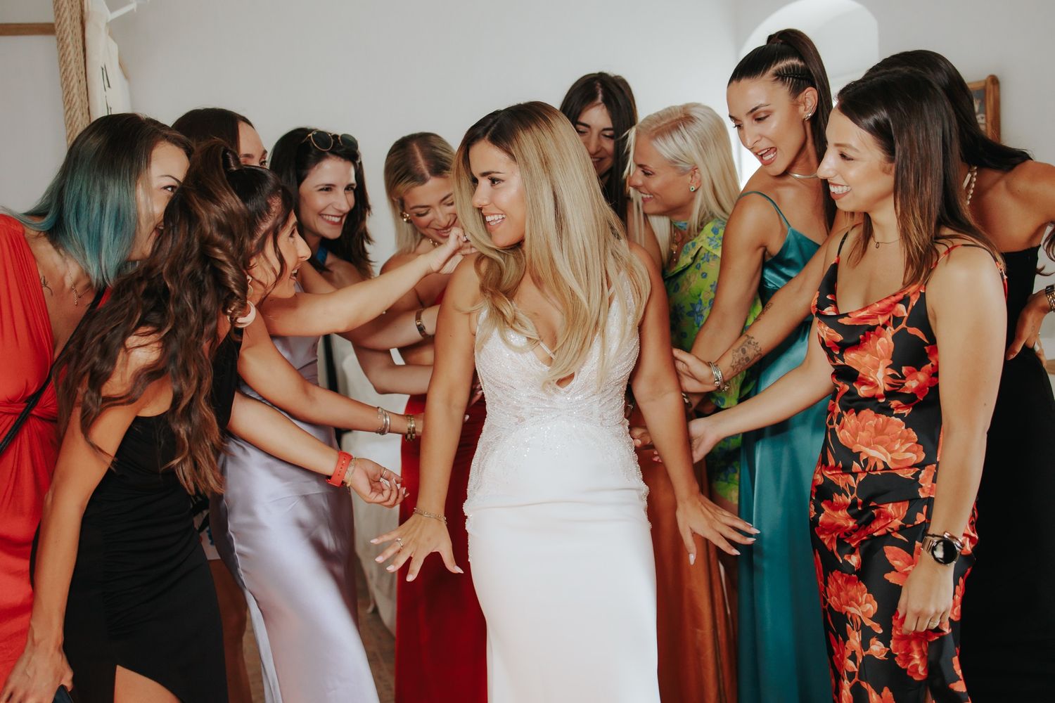 A group of women in colorful dresses assist with final adjustments to a bride's white gown during a wedding celebration.