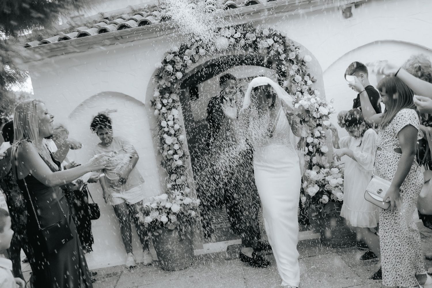 Wedding guests shower rice and confetti on newlyweds under a floral arch in this vintage black and white photograph.