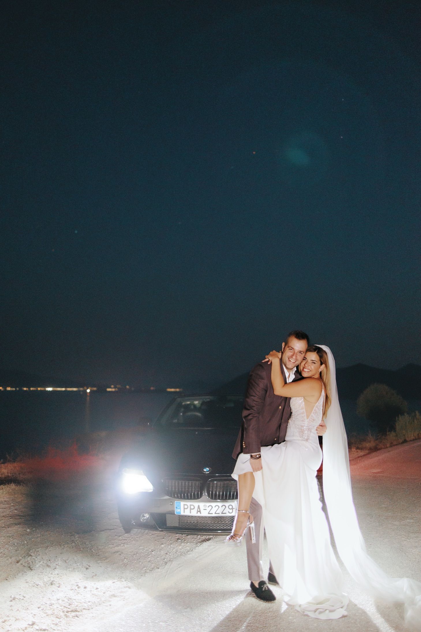 Newlyweds share romantic moment at night with luxury car and headlights illuminating desert landscape.