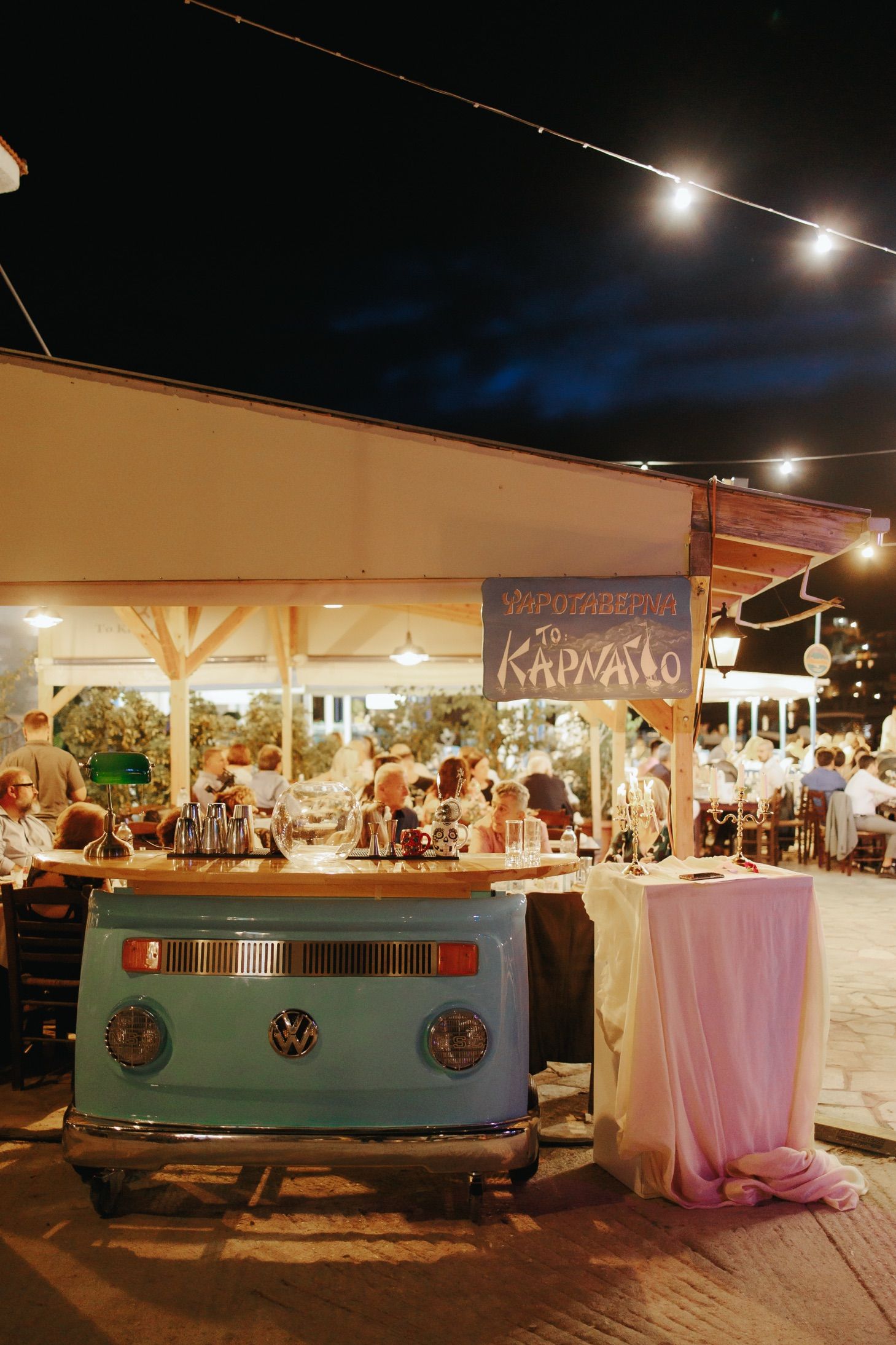 A vintage Volkswagen bus bar setup at an evening outdoor event with string lights and people dining in the background.