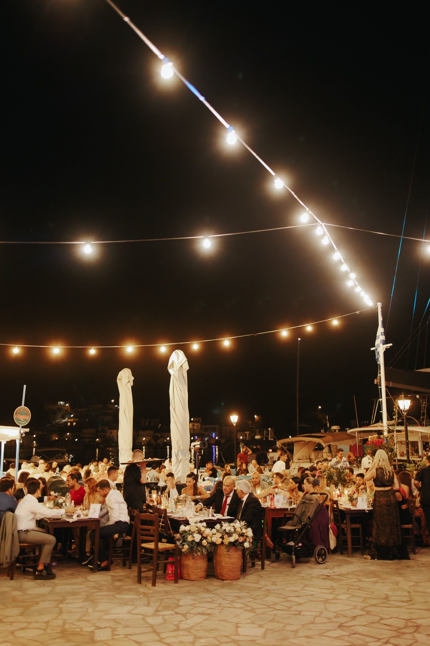 Outdoor evening reception with string lights illuminating tables of guests dining on a stone patio near the waterfront.