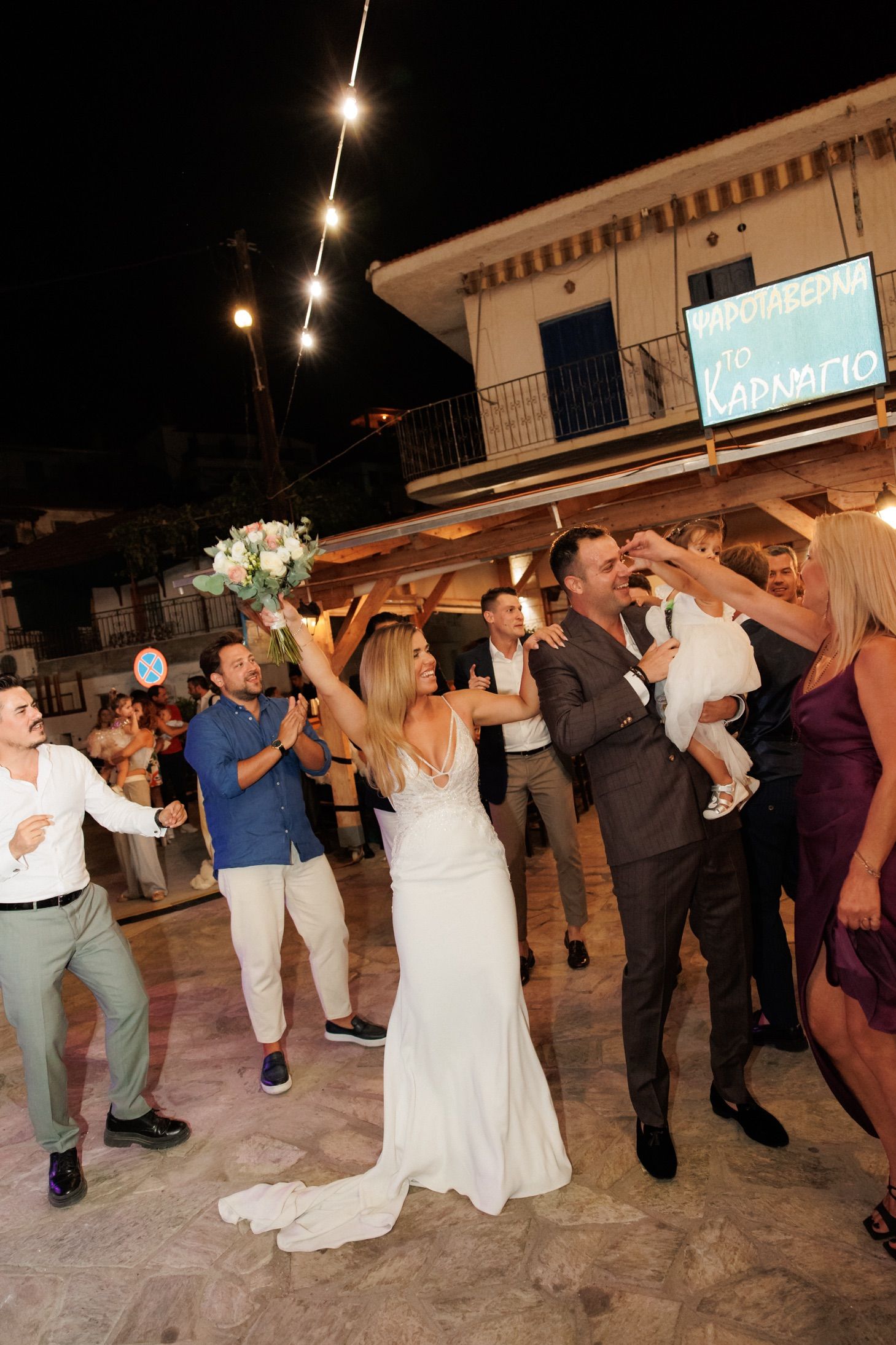 Wedding guests and newlyweds form a dancing line in a festive outdoor reception venue illuminated by string lights.