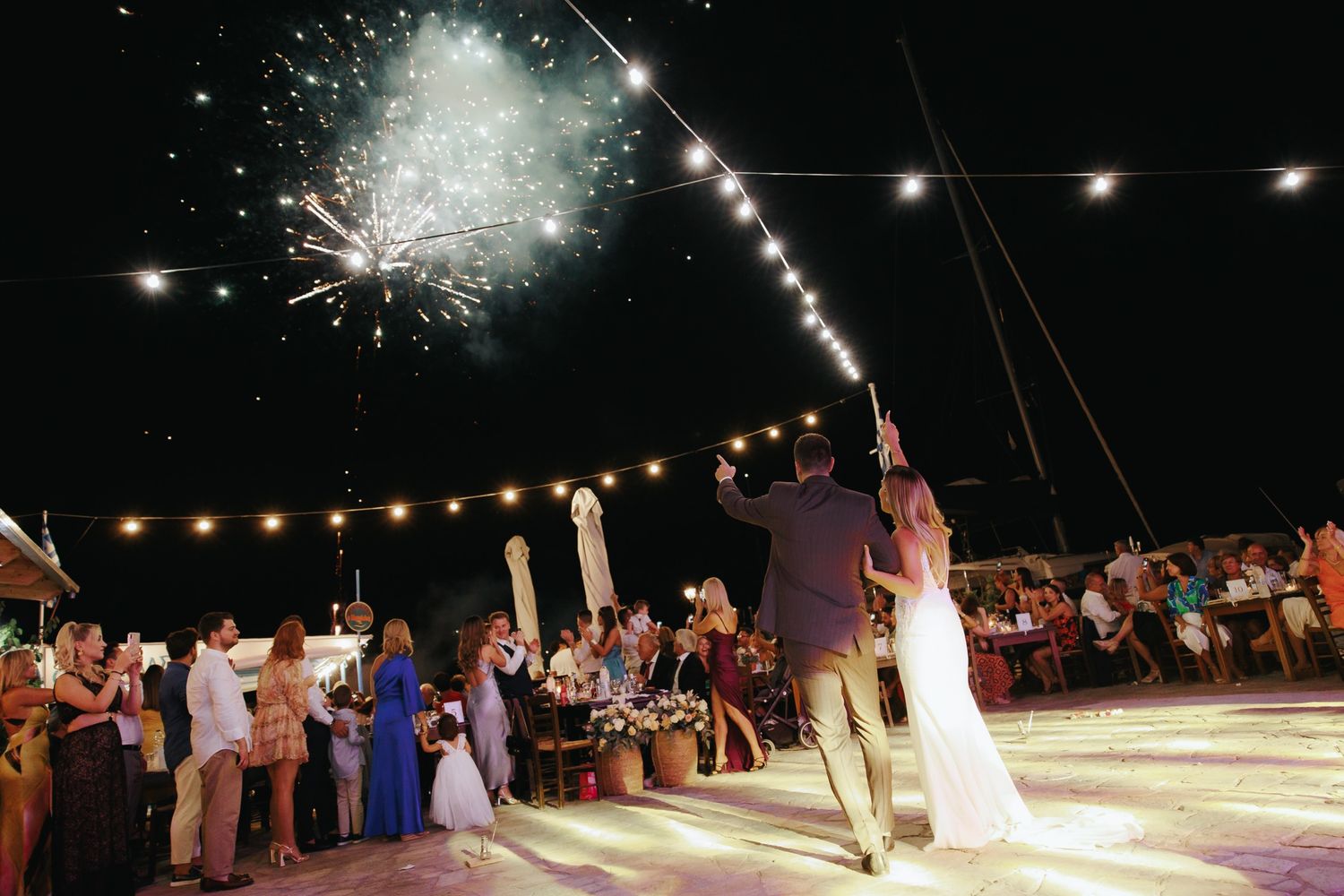 String lights illuminate an outdoor wedding reception dance floor at night as fireworks burst in the night sky.