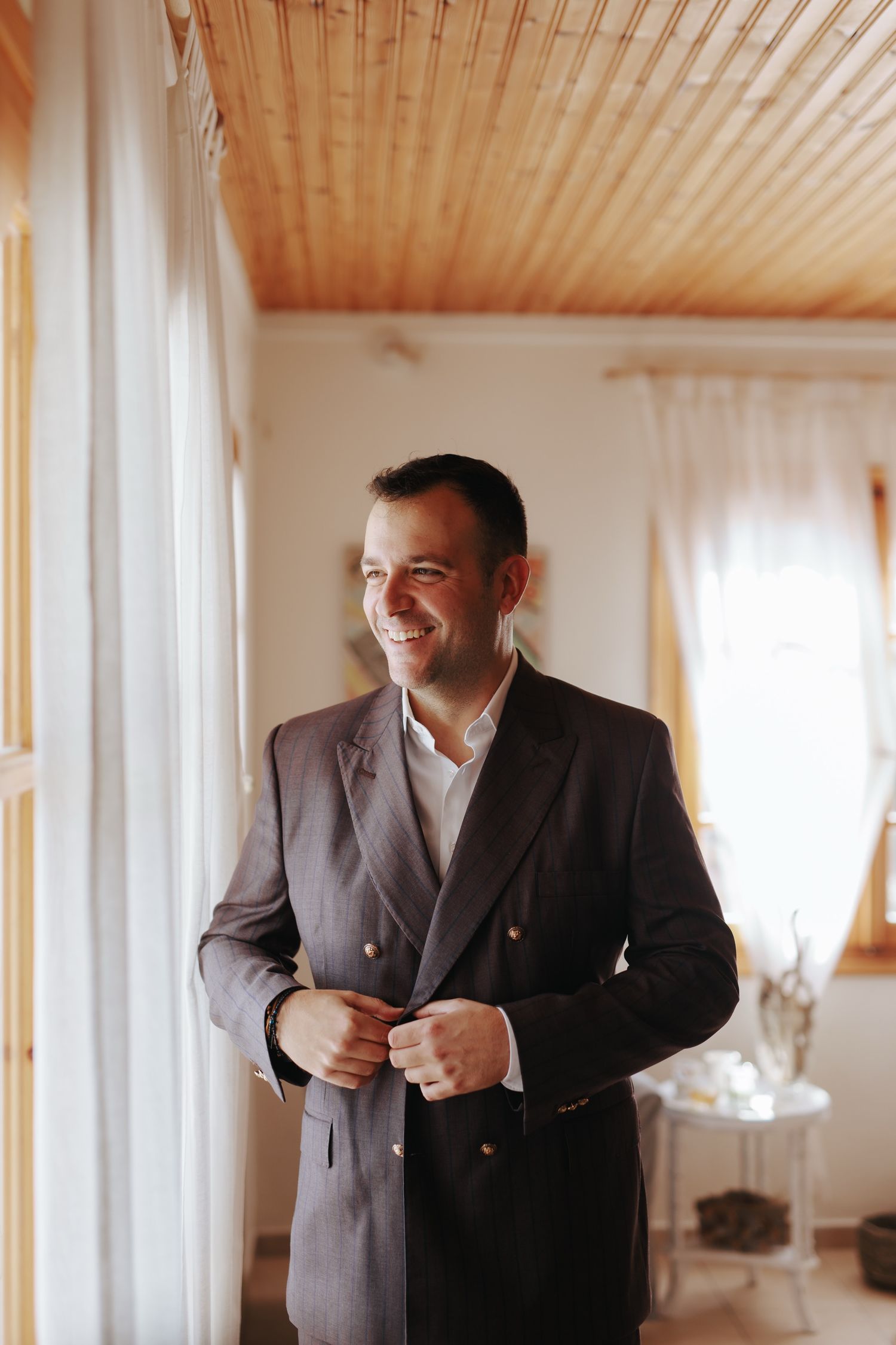A man in a dark suit smiles while adjusting his jacket by a sunlit window.