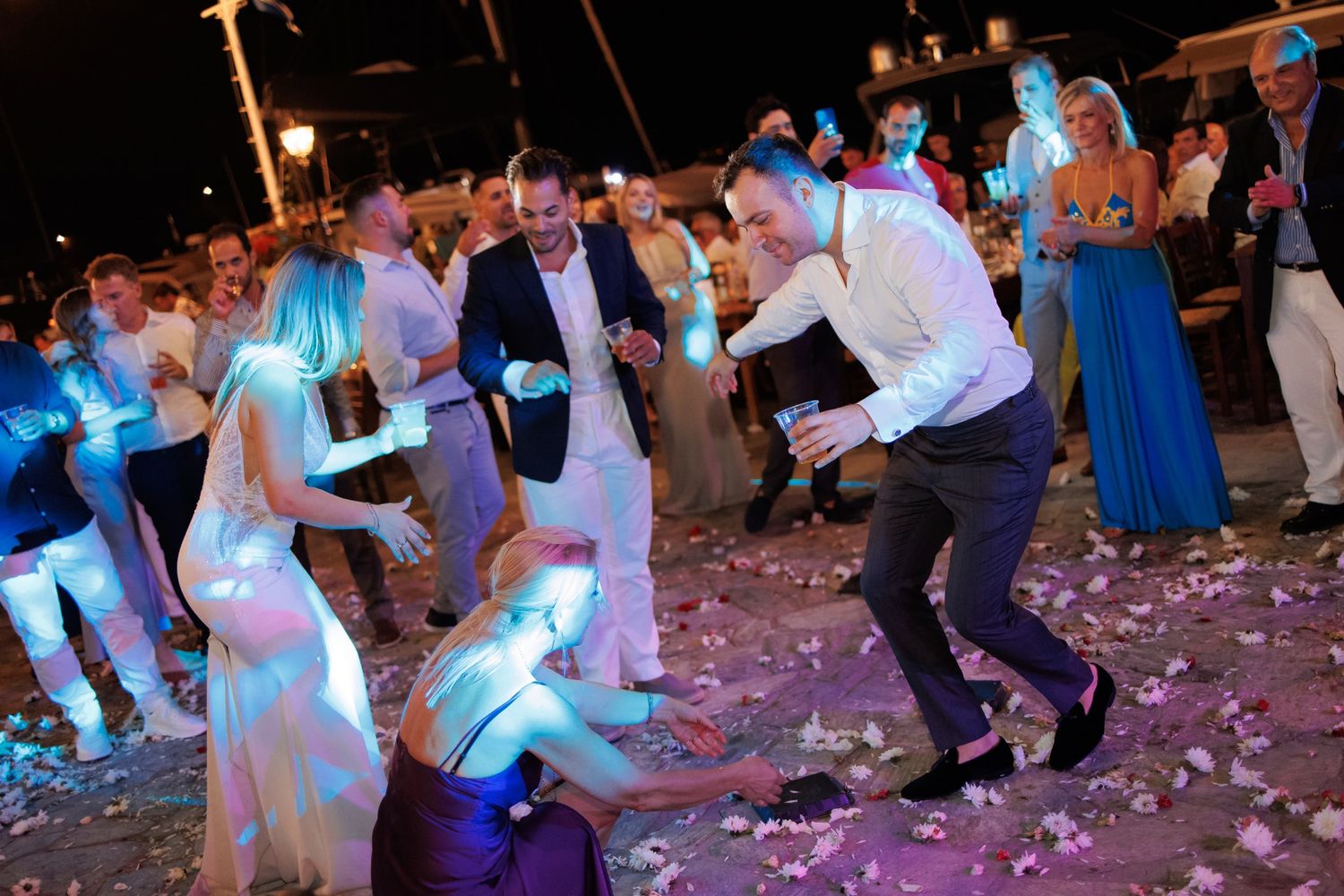 Wedding guests dancing on a confetti-covered dance floor at night with blue lighting and formal attire.