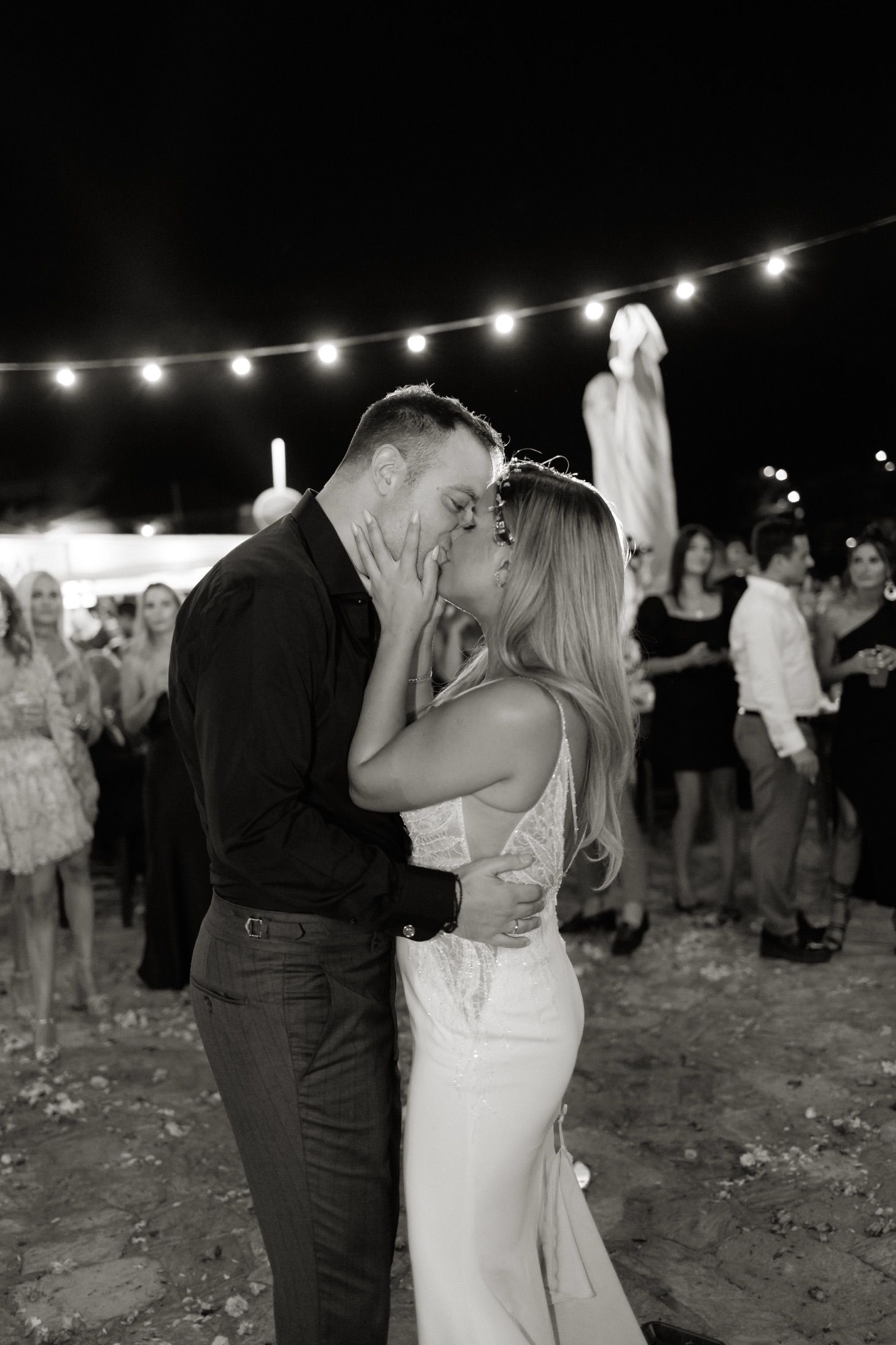 A romantic outdoor wedding moment under string lights with a couple sharing a loving dance at night.