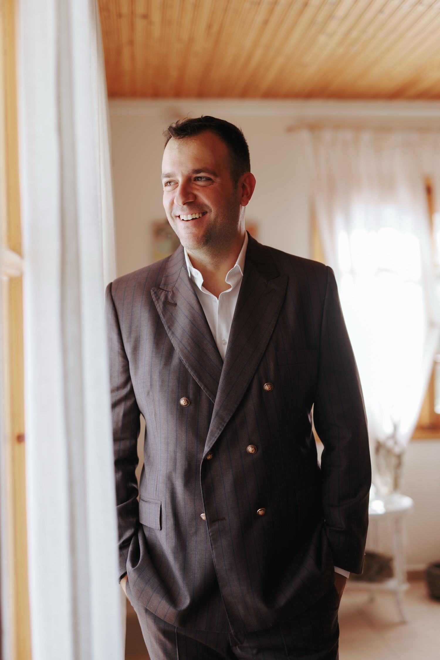 Man in dark business suit smiling while standing in a room with wood paneled ceiling.