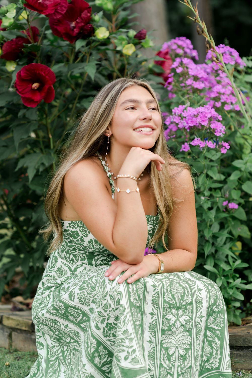 A summer portrait in a garden with deep red hollyhocks and pink phlox flowers framing a green and white patterned dress.