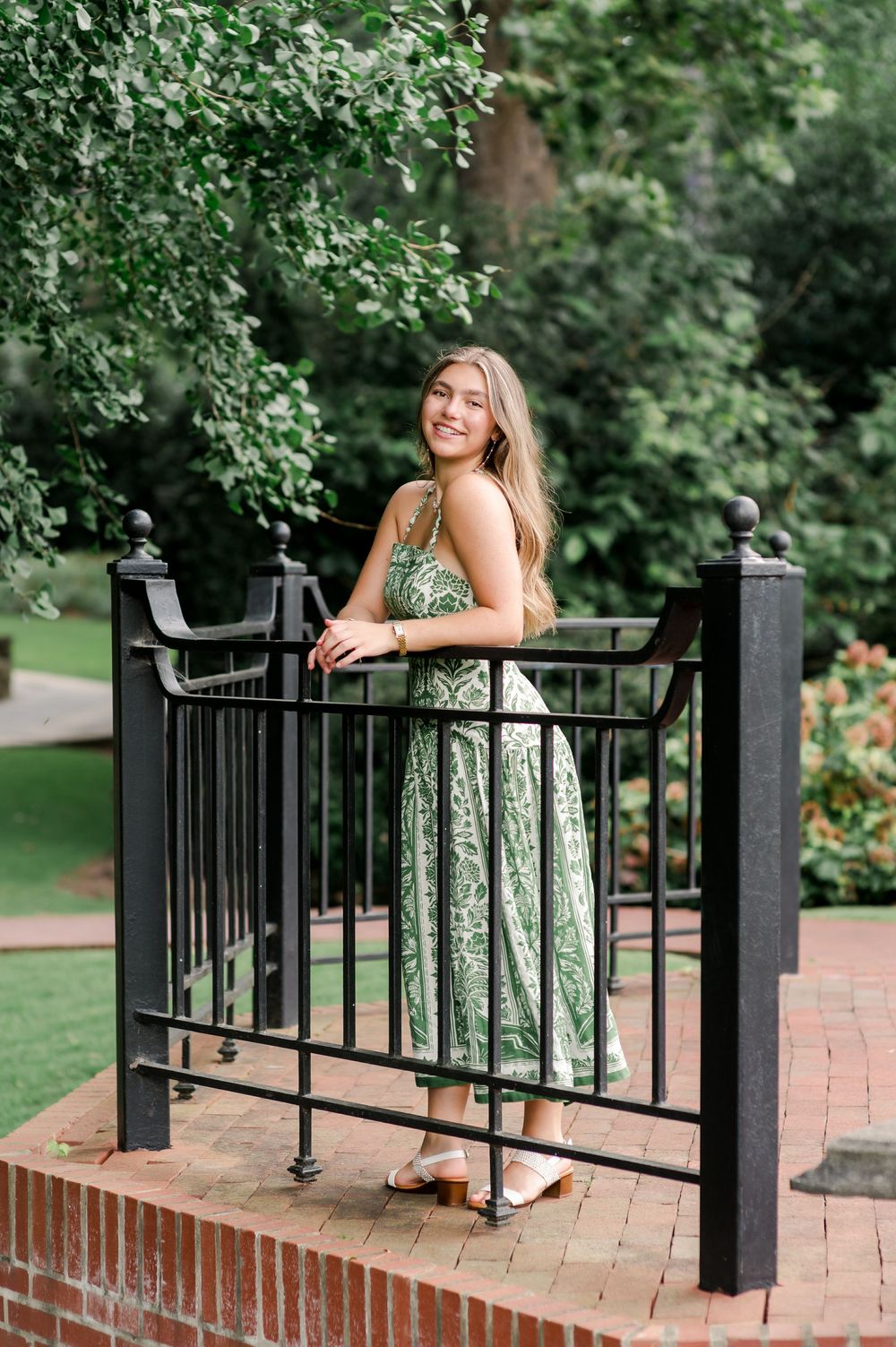 A green floral maxi dress photographed on a brick patio with wrought iron railings and lush garden background.