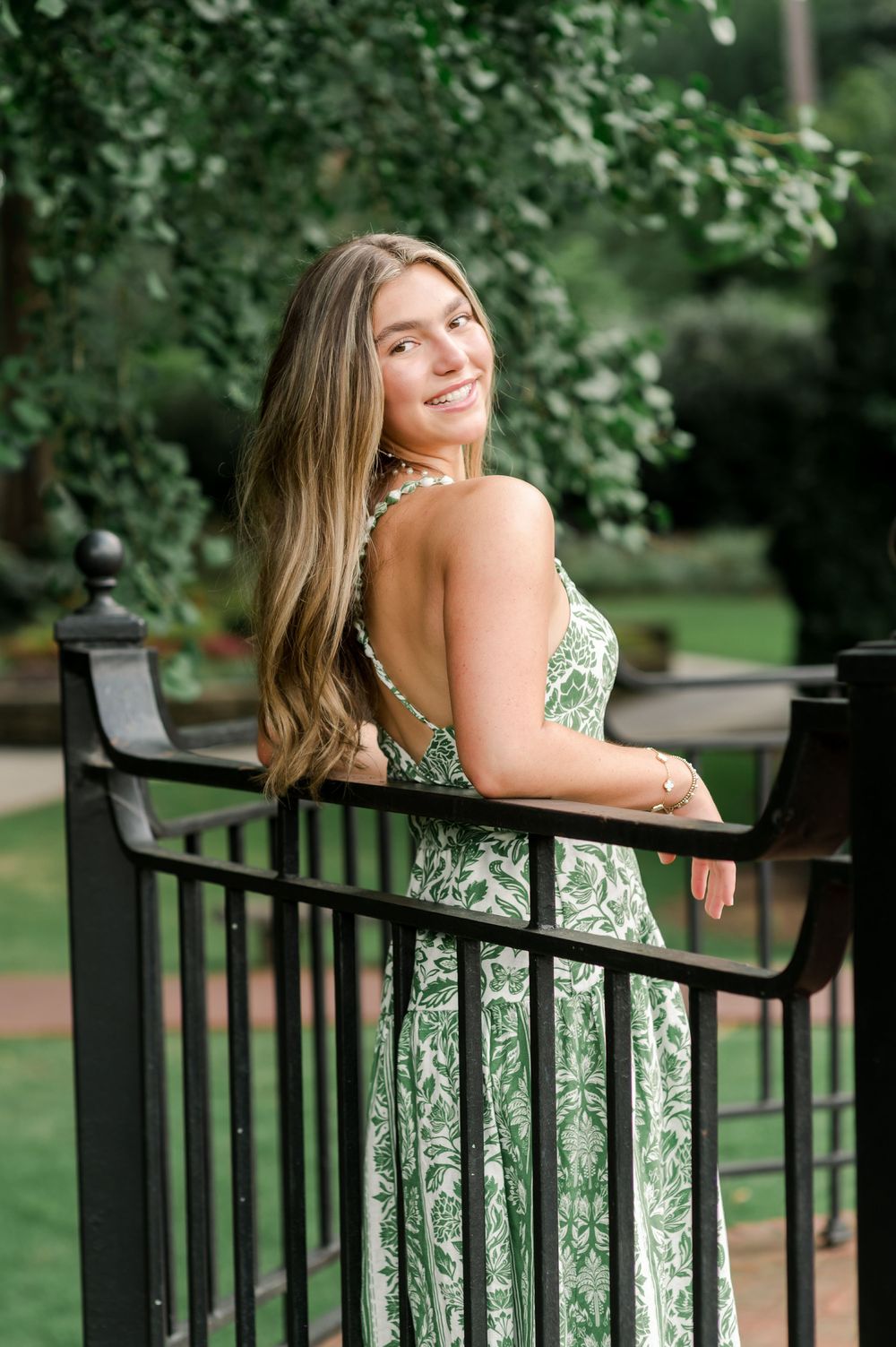A person in a green and white floral sundress poses smiling on a black railing with lush greenery in the background.
