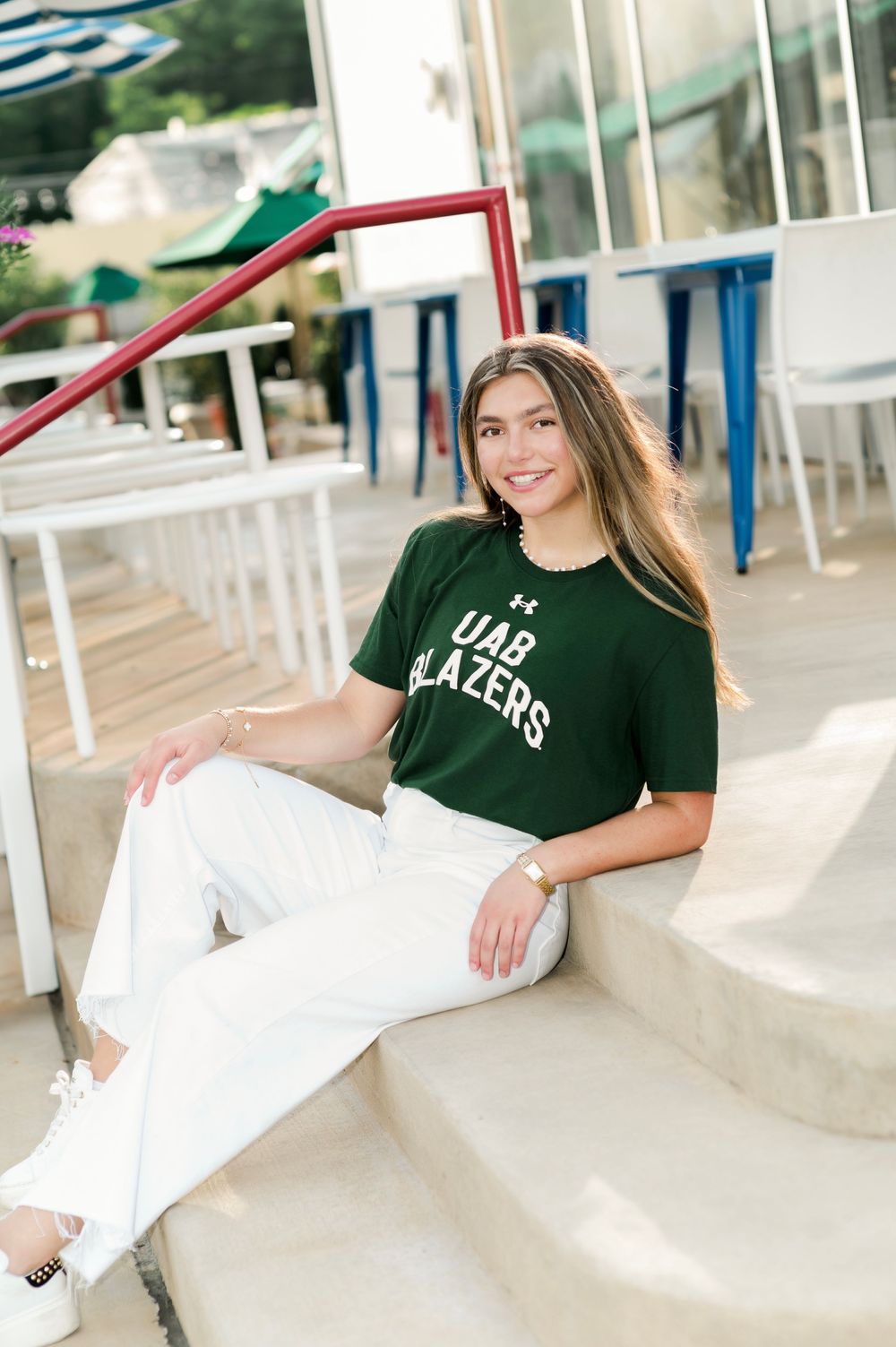 Student wearing a UAB Blazers t-shirt and white pants relaxes on outdoor steps near white and blue seating.