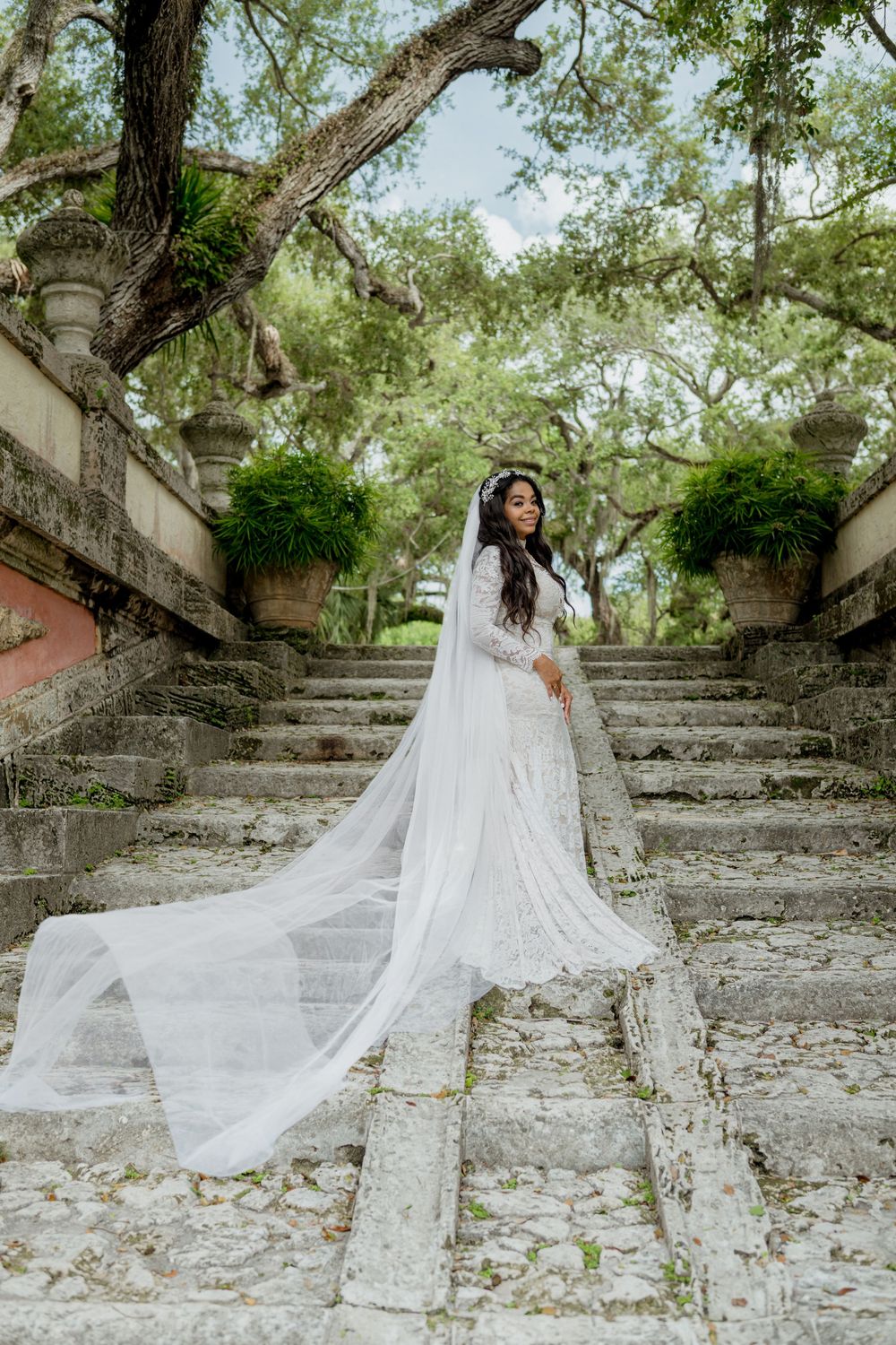 Bride in flowing white lace gown with dramatic long veil poses on stone steps surrounded by lush greenery.