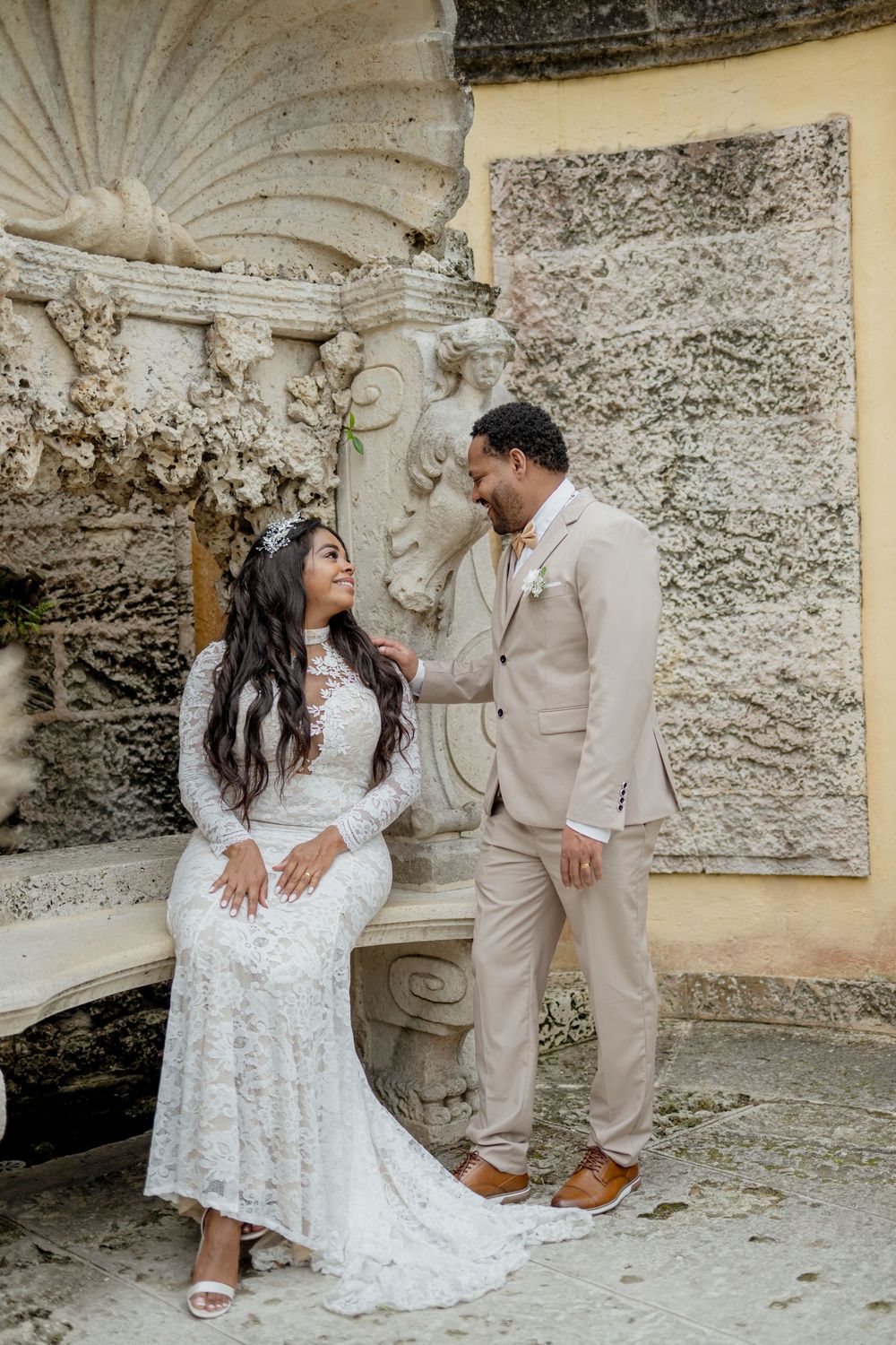 A couple shares a romantic moment in their wedding attire against an ornate stone wall with decorative architectural details.