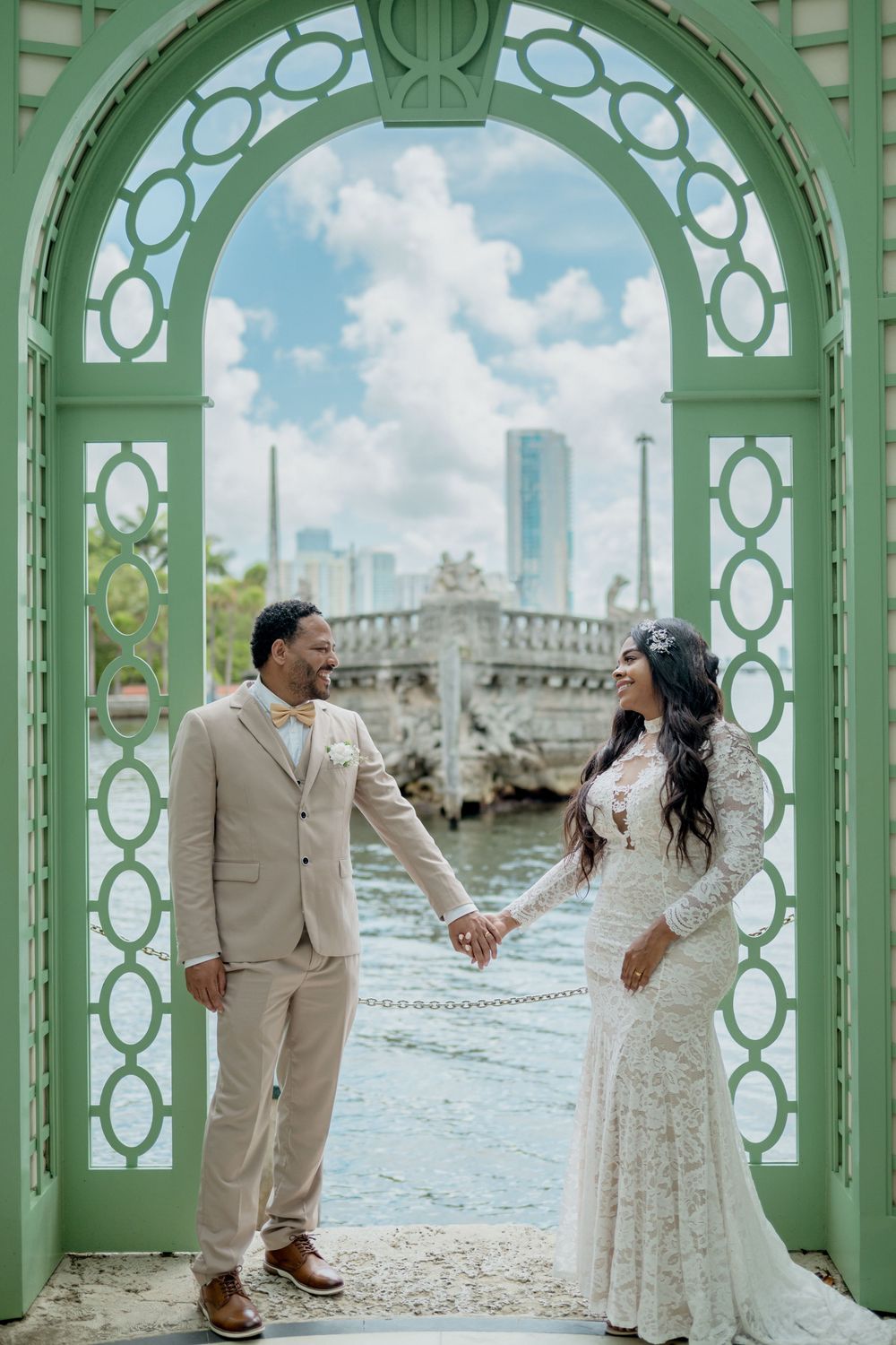 Wedding couple stands under mint green archways by the ocean for romantic beach photos in tropical setting.