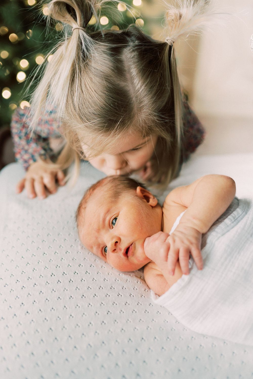 Two siblings share an intimate moment with the older child gazing at the newborn on a white textured surface.