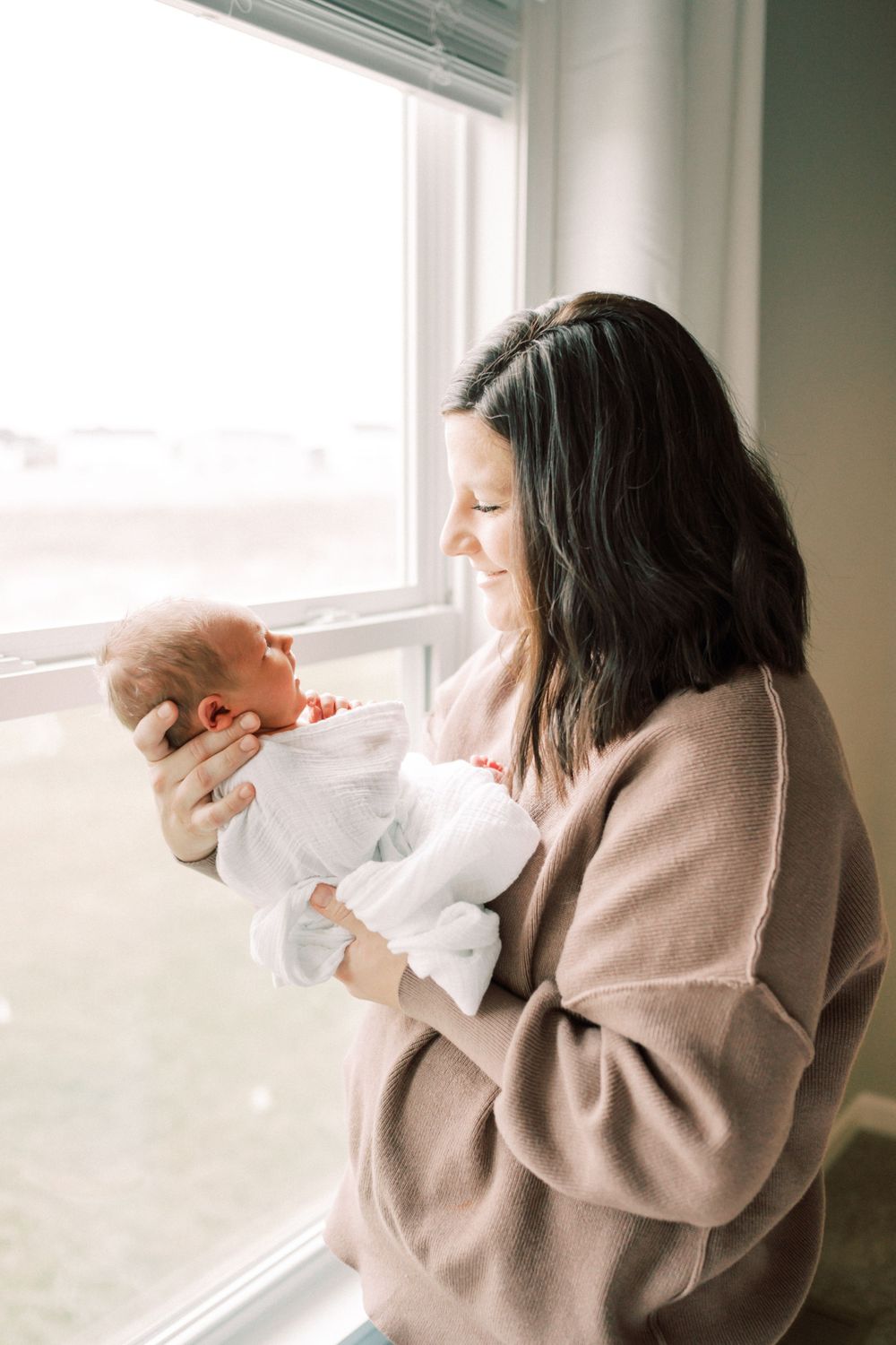 Sweet moment by a bright window as parent cradles newborn wrapped in white.