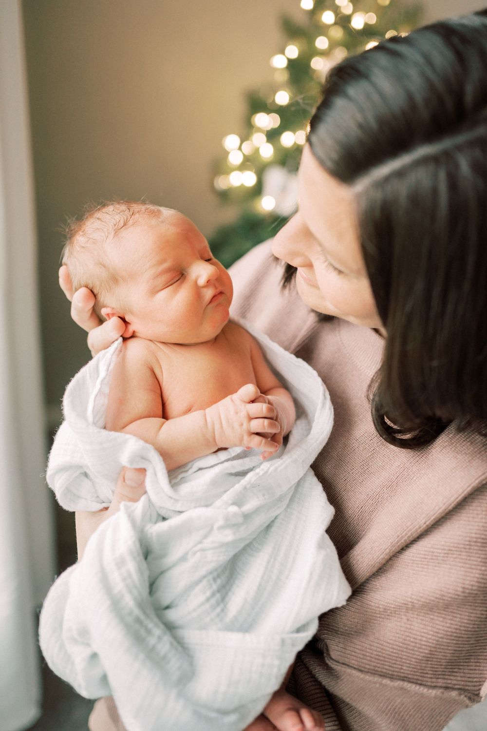 Tender moment with newborn baby wrapped in white near a decorated Christmas tree.