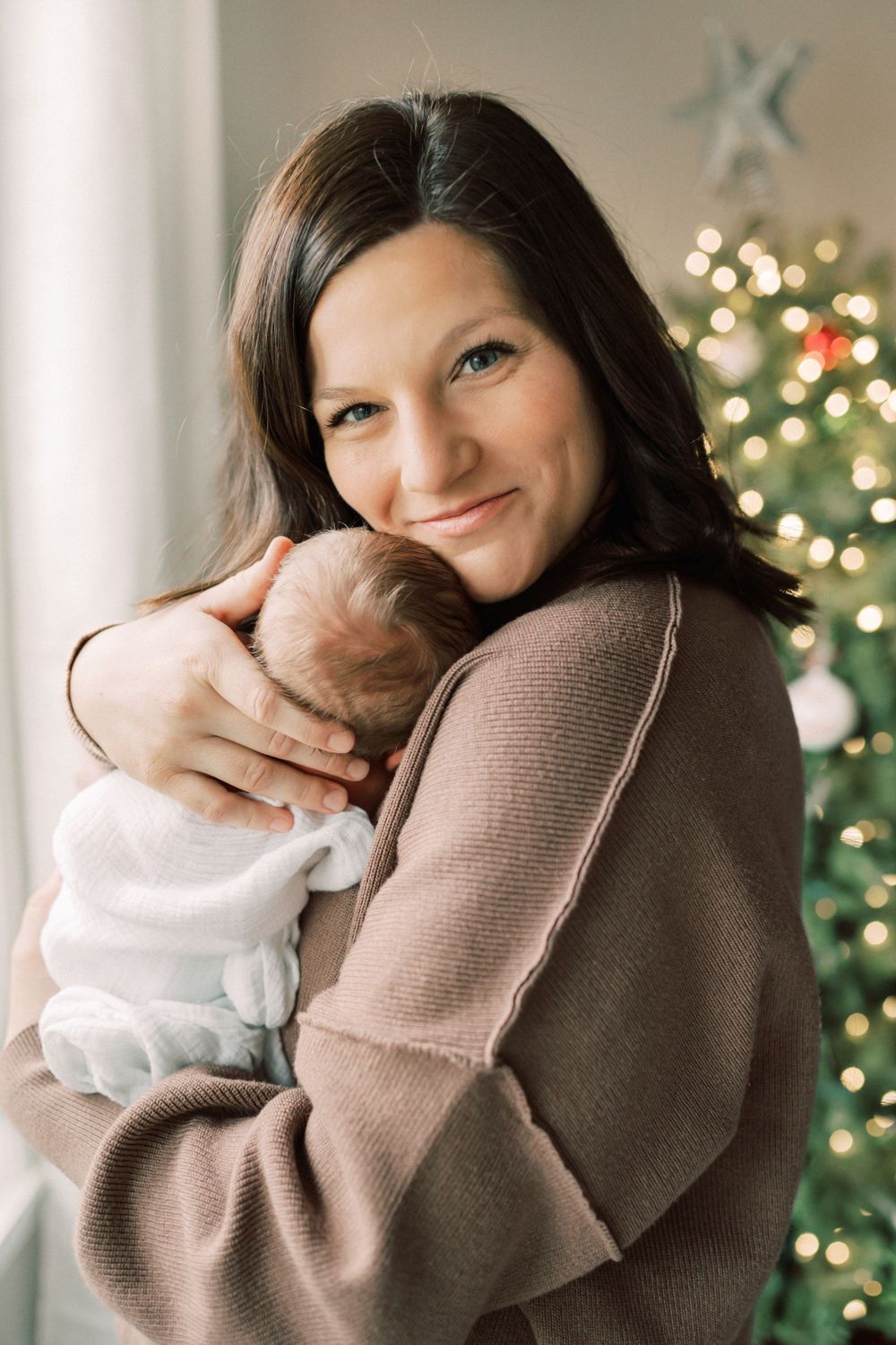 Cozy winter portrait with sleeping newborn against Christmas tree backdrop.