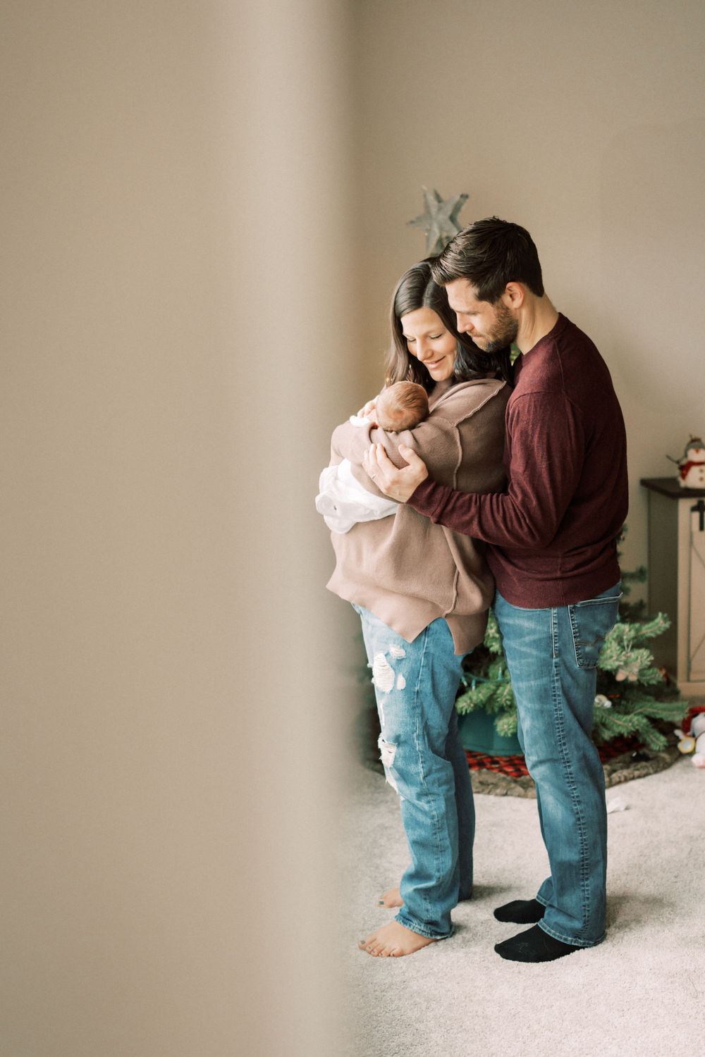 A tender family moment showing parents embracing their newborn baby while standing against a neutral wall.
