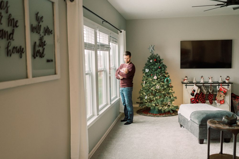 Living room decorated for Christmas with a decorated tree and someone standing by large windows.