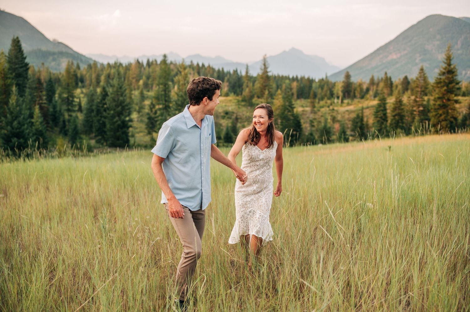 Two people share a joyful moment while standing in a tall grass meadow with mountains and forest in the background.