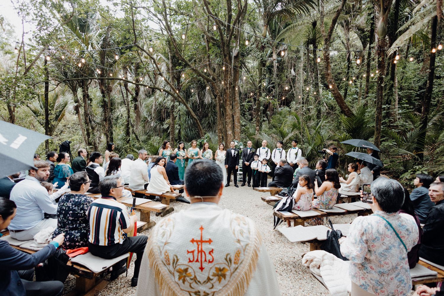 An outdoor Catholic Wedding Ceremony with guests seated on wooden benches captured by Apertured Media Auckland Wedding Photographer.