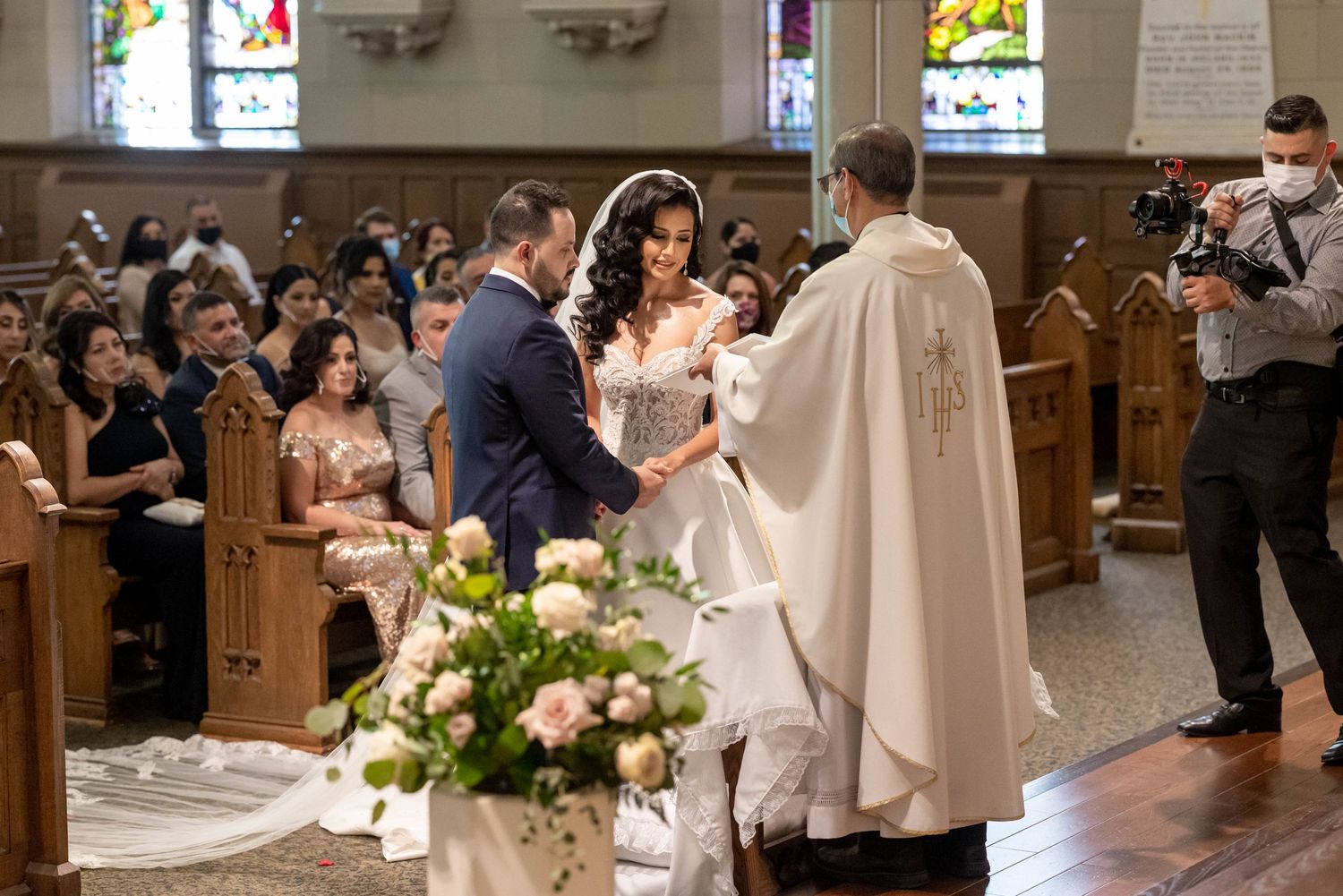 A traditional Catholic wedding ceremony takes place in a church with floral arrangements and stained glass windows in view.