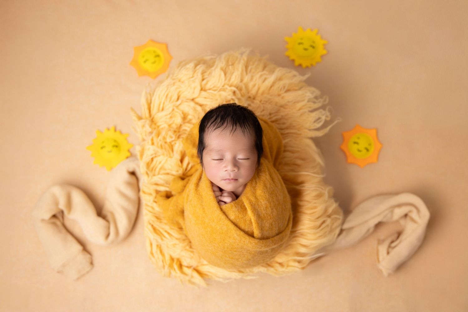 Sleeping swaddled newborn on beige background surrounded by yellow flowers and fuzzy decorative elements.