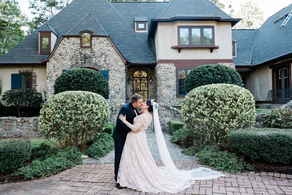 A romantic moment between newlyweds in front of a stone and brick mansion with manicured shrubs and a brick pathway.