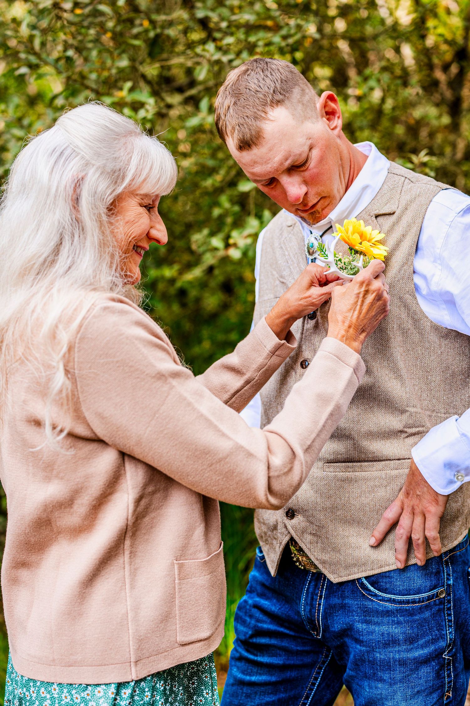 A woman in a beige sweater pins a yellow sunflower boutonniere onto someone's vest at an outdoor event.