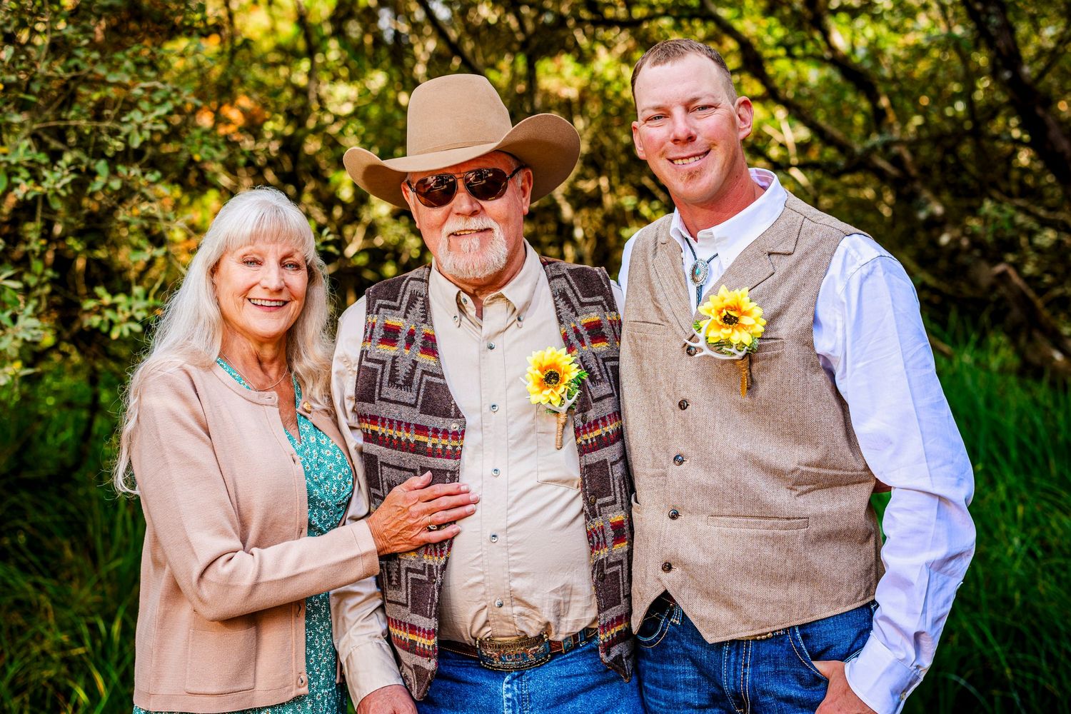 Three people in casual western attire standing close together outdoors with yellow flower boutonnieres and sunlit foliage.